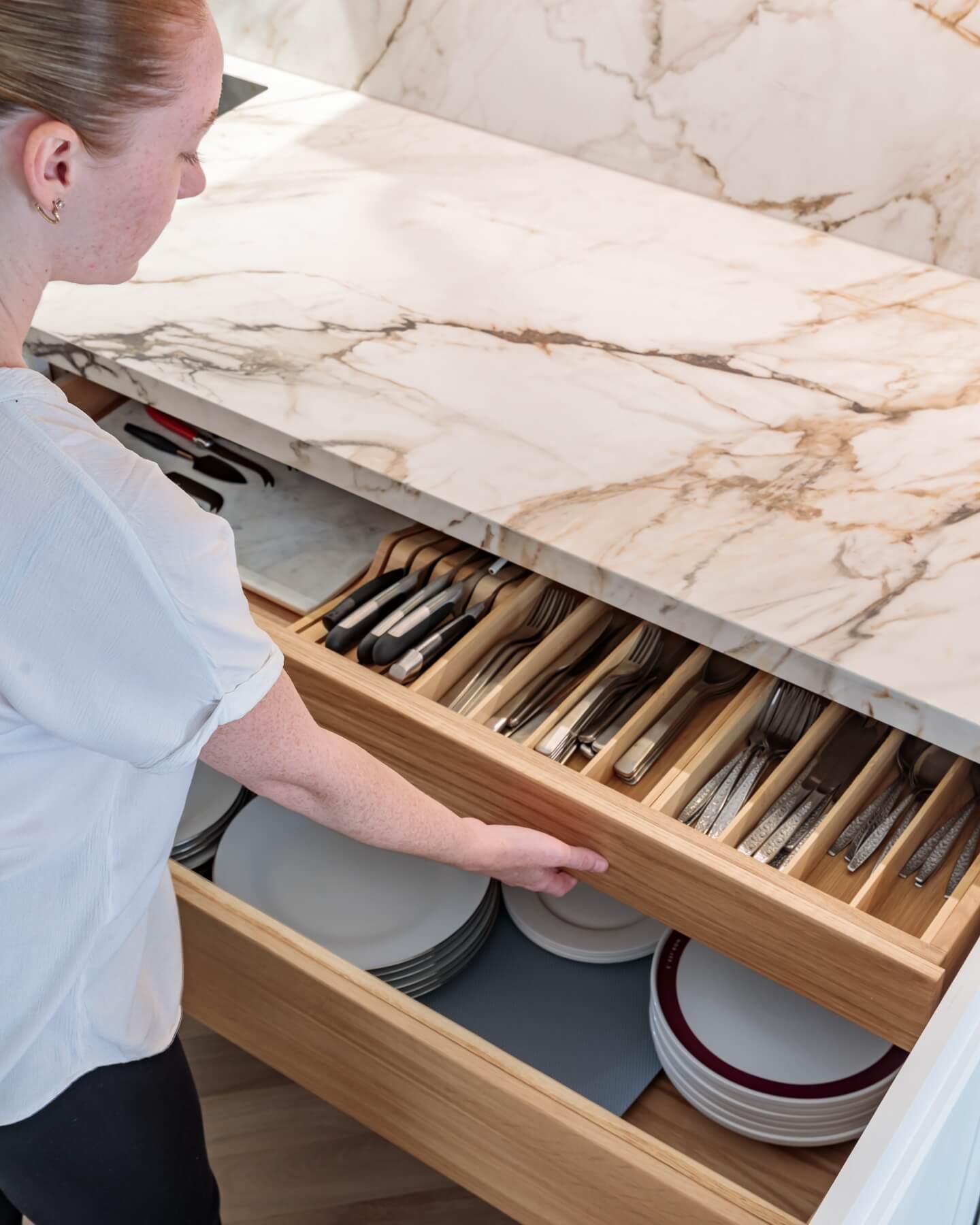 Arch KBB team member organising cutlery in a wooden drawer beneath a marble countertop, with neatly arranged utensils and plates visible in the background.