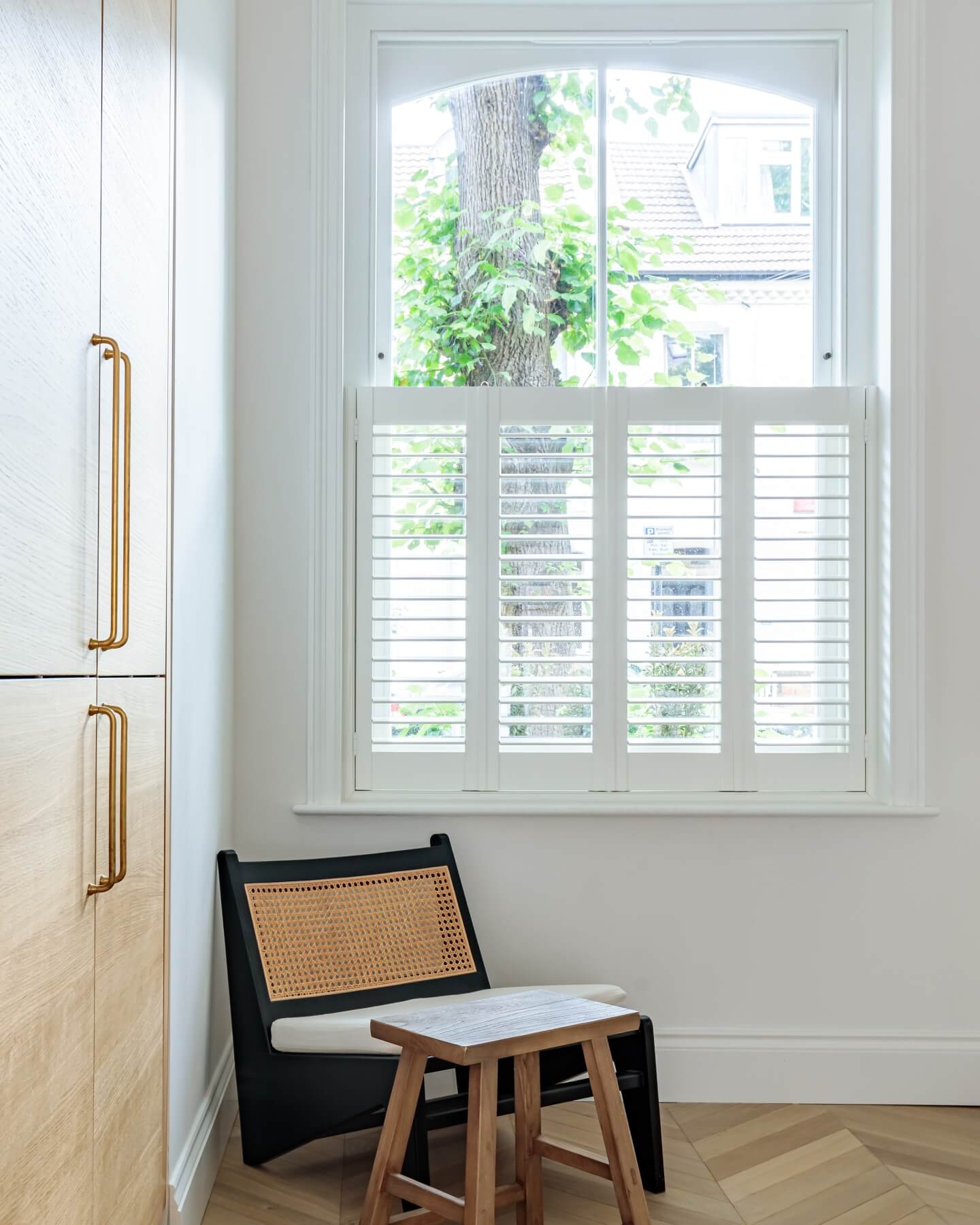 Bright interior space featuring a modern window with white shutters, allowing natural light to filter in. A stylish black chair with a woven seat is positioned beside a wooden cabinet with gold handles, creating a contemporary and inviting atmosphere. An Arch KBB team member is present, contributing to the design and decor.