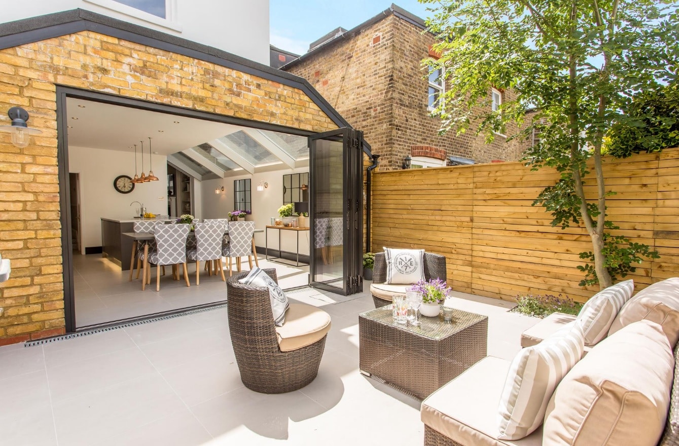 Outdoor living space featuring a contemporary patio with seating, a glass table adorned with flowers, and an open doorway leading to a stylish kitchen area. The Arch KBB team member is present, showcasing the seamless indoor-outdoor design. Surrounding elements include wooden fencing and greenery, enhancing the inviting atmosphere.