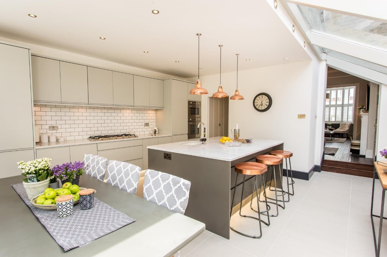 Modern kitchen interior featuring an Arch KBB team member, with sleek cabinetry, a spacious island, and stylish copper pendant lighting. The design includes a dining area with patterned chairs and a bowl of fresh fruit, creating a warm and inviting atmosphere.