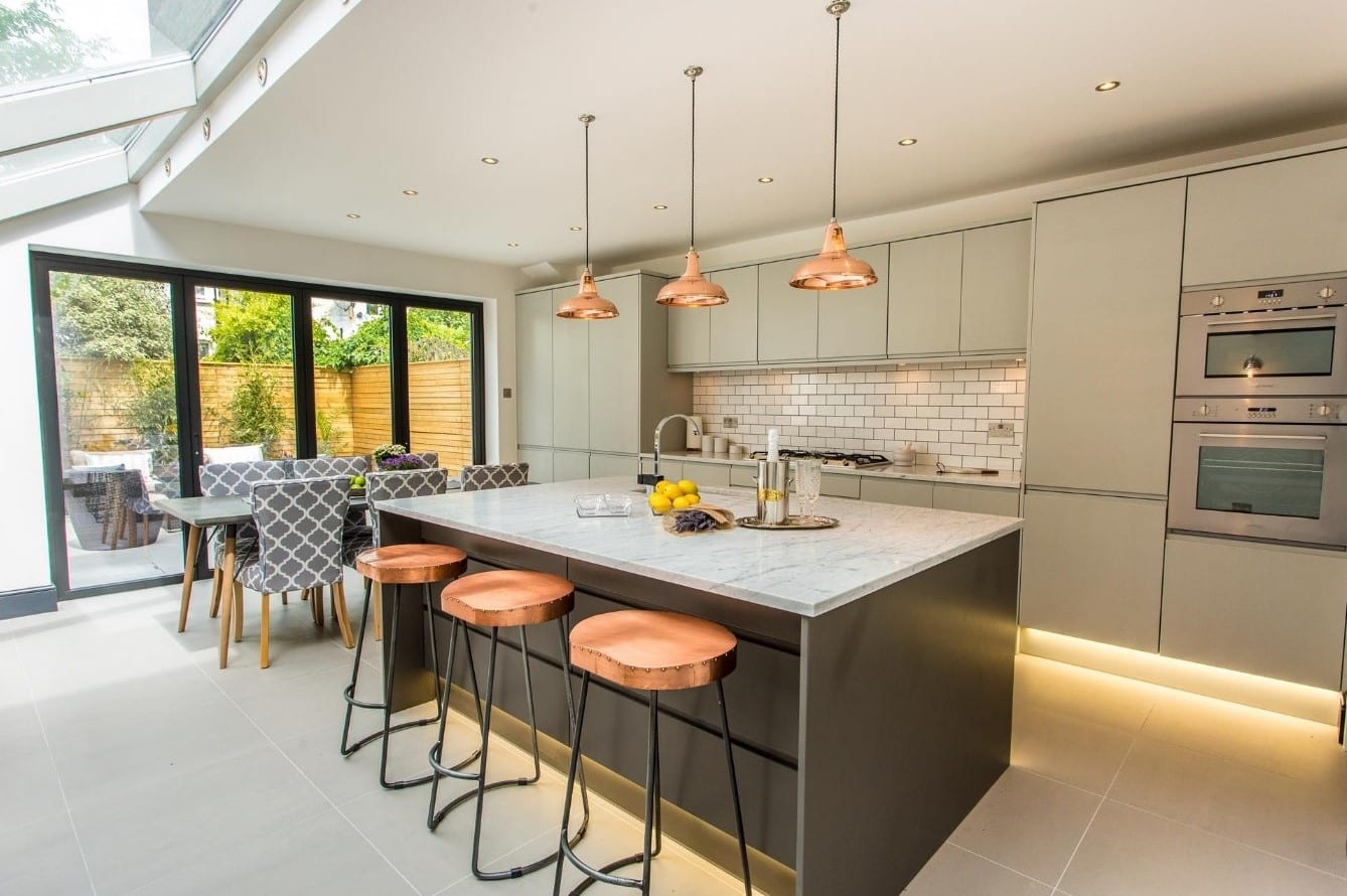 Modern kitchen featuring a large island with copper bar stools, pendant lighting, and sleek cabinetry. The space includes a stylish dining area with patterned chairs and large glass doors leading to an outdoor patio, showcasing a bright and inviting atmosphere. An Arch KBB team member is present, enhancing the kitchen's appeal.