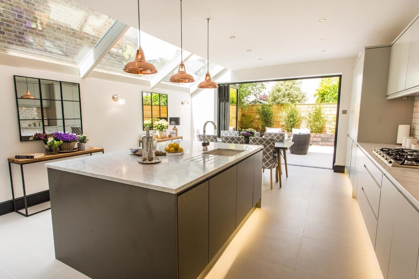 Modern kitchen featuring a central island with a marble countertop, sleek cabinetry, and copper pendant lights. The space is illuminated by natural light streaming through skylights, with an open view to a garden area. An Arch KBB team member can be seen in the kitchen, enhancing the contemporary design.