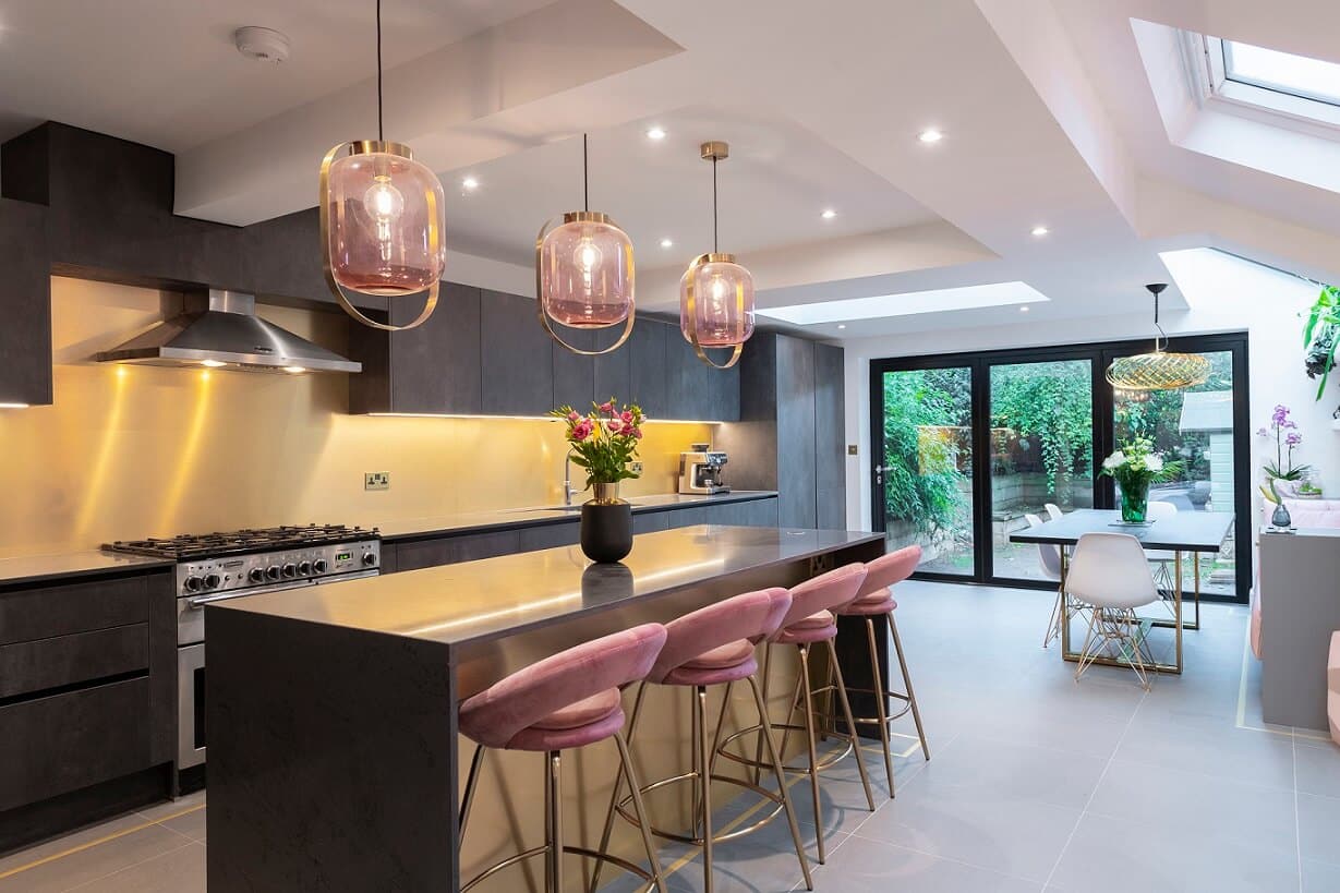 Modern kitchen design featuring a sleek island with pink bar stools, stylish pendant lighting, and a spacious layout. The Arch KBB team member is present, showcasing the contemporary cabinetry and appliances, with a view of the garden through large glass doors. Ideal for inspiration in home renovations.