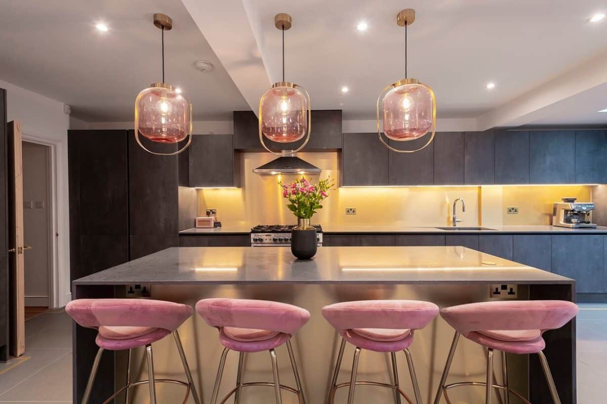 Modern kitchen featuring a sleek island with a grey countertop, four stylish pink bar stools, and elegant pendant lighting. The space is enhanced by warm ambient lighting and a floral arrangement in a black vase, creating a welcoming atmosphere. An Arch KBB team member is present, showcasing the kitchen's design and functionality.