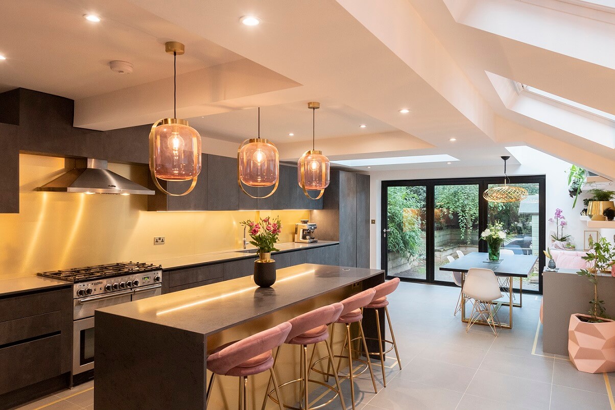 Modern kitchen design featuring a spacious island with pink bar stools, stylish pendant lighting, and sleek cabinetry. An Arch KBB team member is present, enhancing the aesthetic of the contemporary space that opens to a garden area through large glass doors.