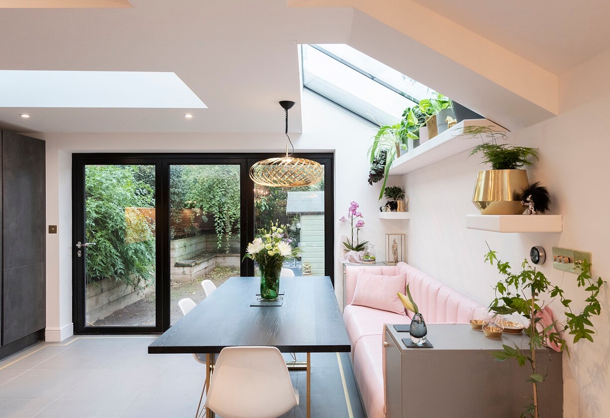 Bright and modern kitchen-diner featuring a sleek black dining table, white chairs, and a pink sofa. Large glass doors open to a lush garden, while a skylight provides ample natural light. An Arch KBB team member is present, adding to the welcoming atmosphere. Decorative plants and stylish accessories enhance the contemporary design.