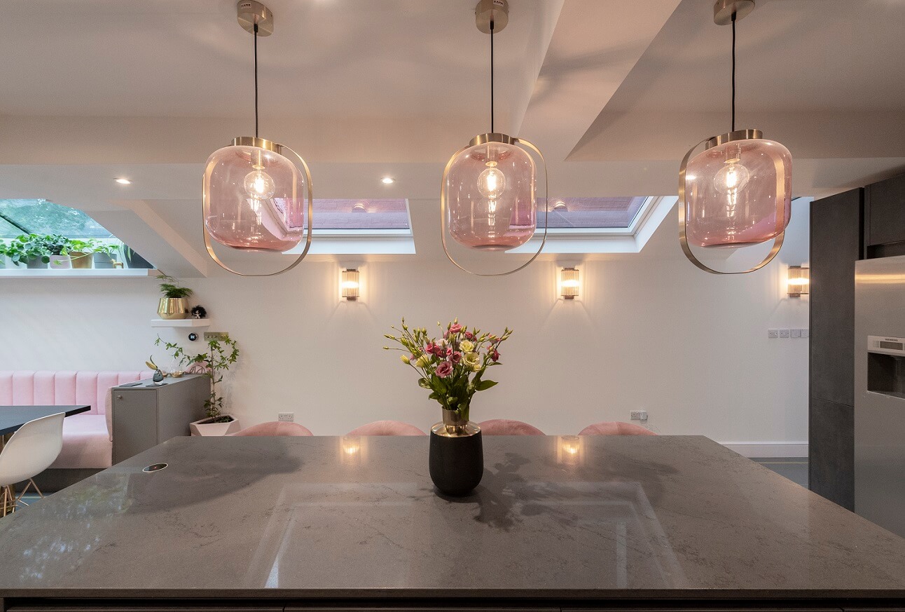 Modern kitchen interior featuring a sleek grey worktop, a vase of fresh flowers, and stylish pendant lighting. The space is illuminated by natural light from skylights, complemented by soft pink seating and contemporary wall lighting. An Arch KBB team member is present, contributing to the inviting atmosphere.