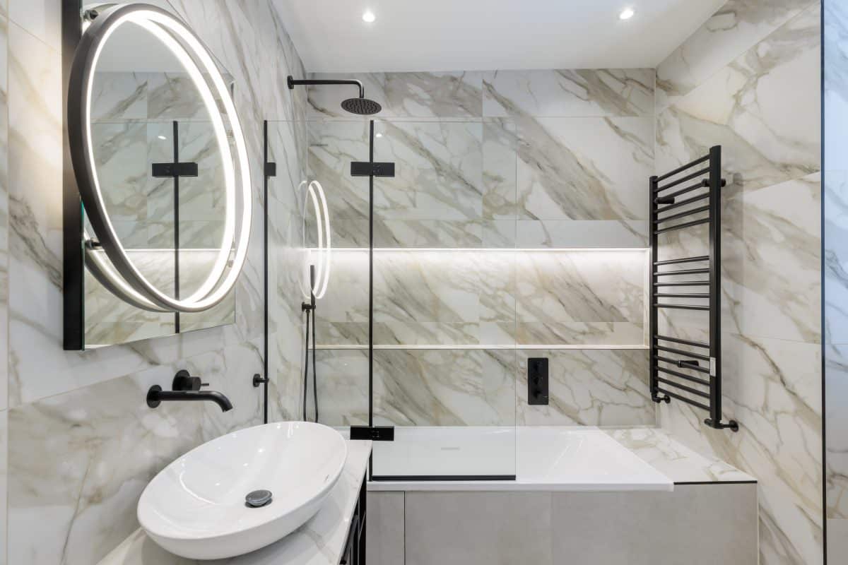 Modern bathroom design featuring elegant marble wall tiles, a sleek circular mirror, a stylish wash basin, and a contemporary shower area. The space is complemented by a black heated towel rail and ambient lighting, showcasing a blend of functionality and luxury. An Arch KBB team member is present, enhancing the professional atmosphere.