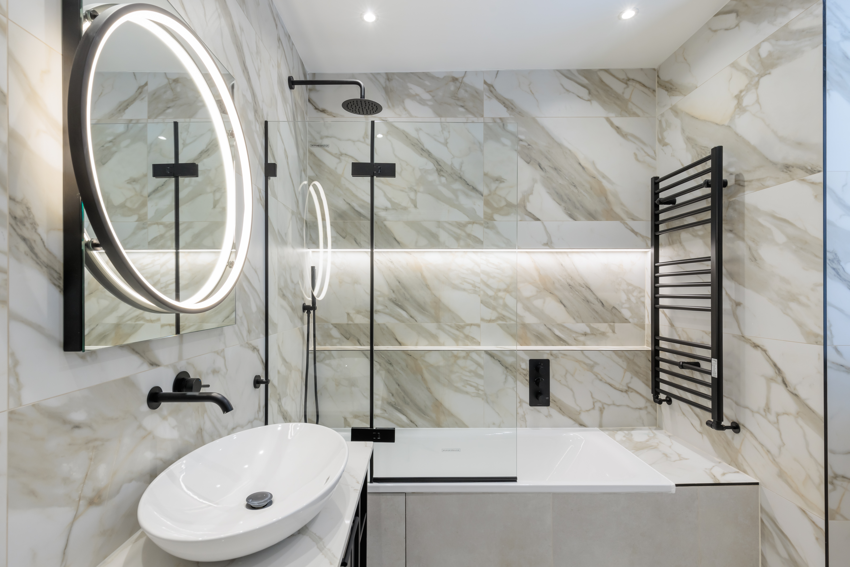 Modern bathroom design featuring elegant marble wall tiles, a sleek circular mirror, a stylish wash basin, and a contemporary shower area. The space is complemented by a black heated towel rail and ambient lighting, showcasing a blend of functionality and luxury. An Arch KBB team member is present, enhancing the professional atmosphere.