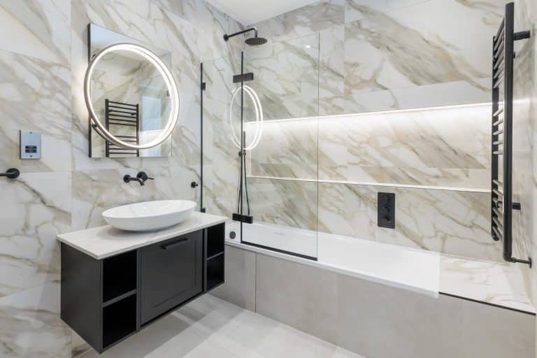 Modern bathroom featuring elegant marble walls, a stylish round mirror with integrated lighting, a sleek basin on a vanity unit, and a glass shower enclosure. An Arch KBB team member is present, showcasing contemporary design elements and functional space.