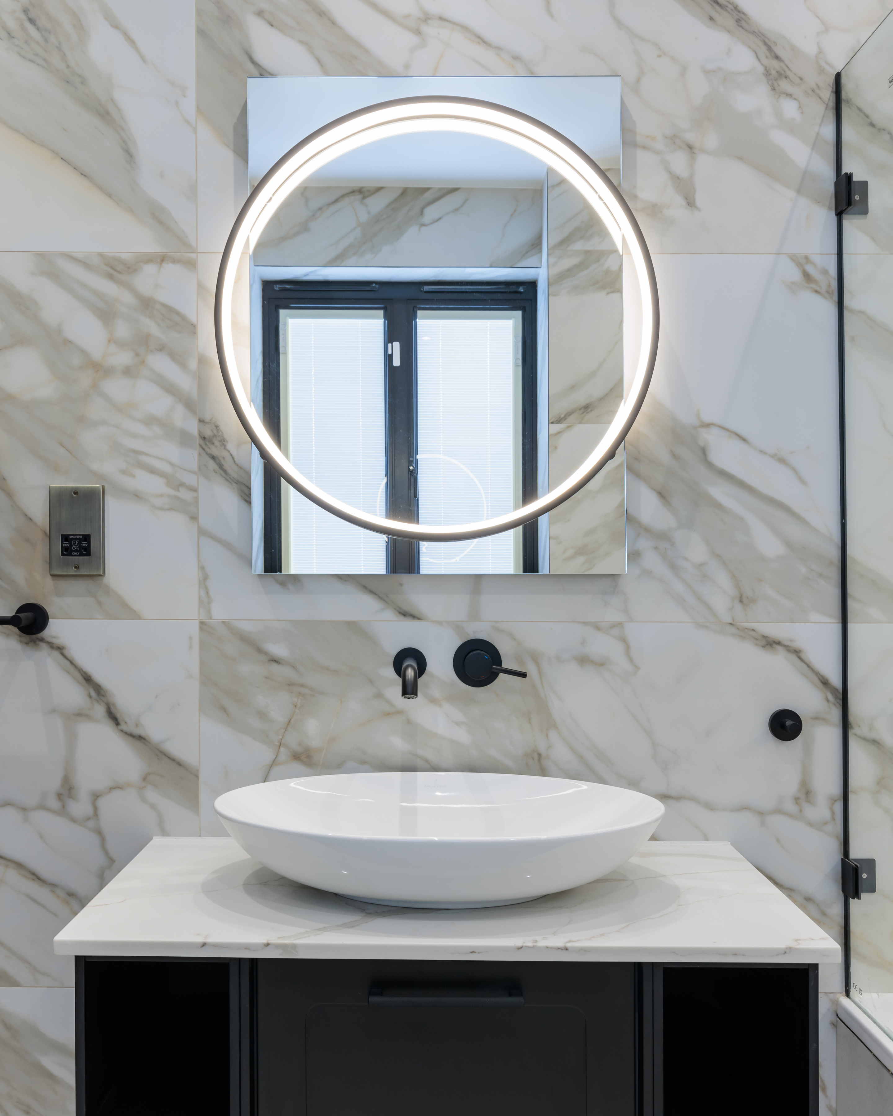 Modern bathroom design featuring a sleek round mirror with integrated lighting, a minimalist white basin, and stylish black fixtures against a marble wall. An Arch KBB team member is present, showcasing contemporary interior aesthetics.