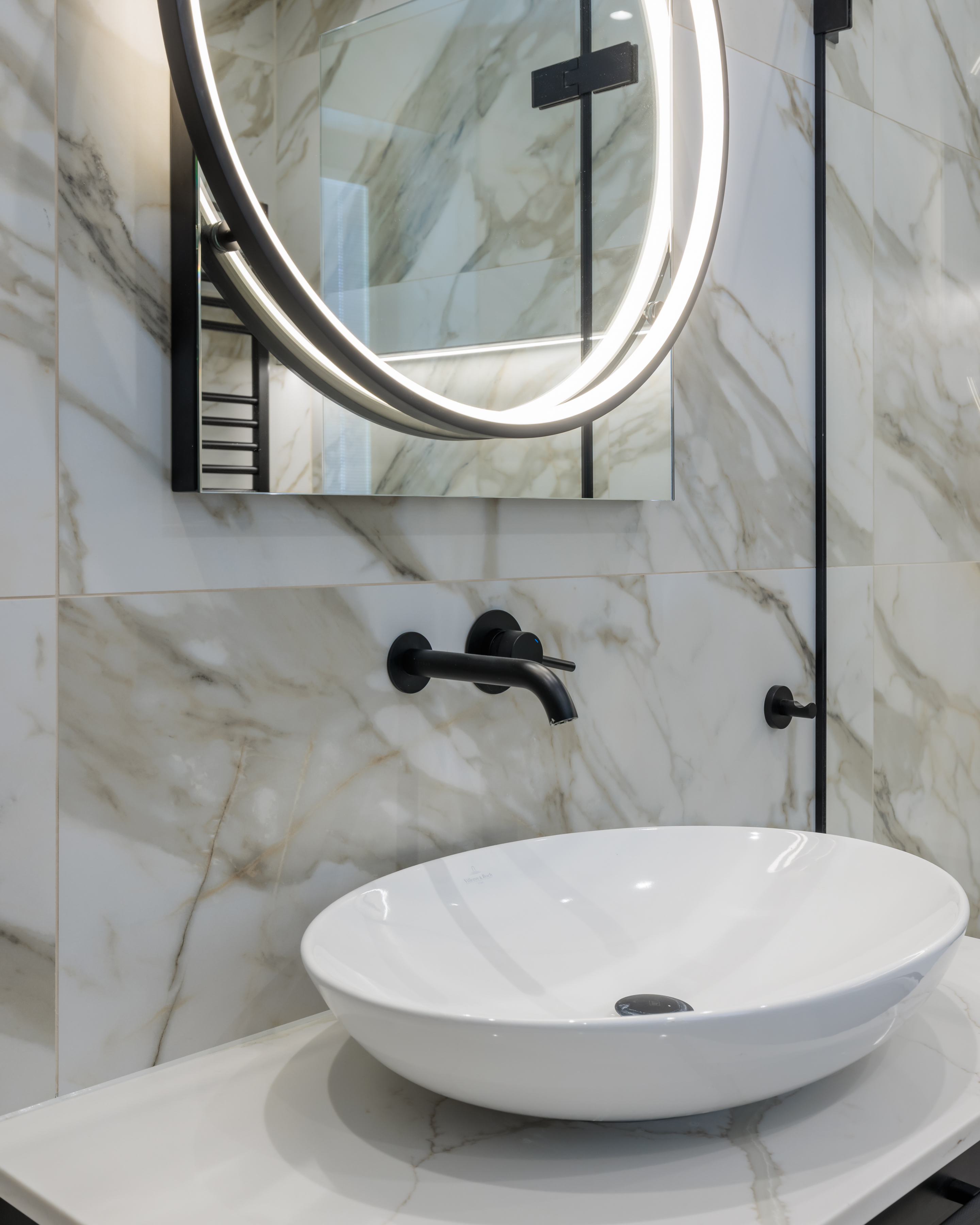 Modern bathroom design featuring a sleek black tap and a round illuminated mirror above a white basin, set against elegant marble wall tiles. An Arch KBB team member is present, showcasing the stylish interior.