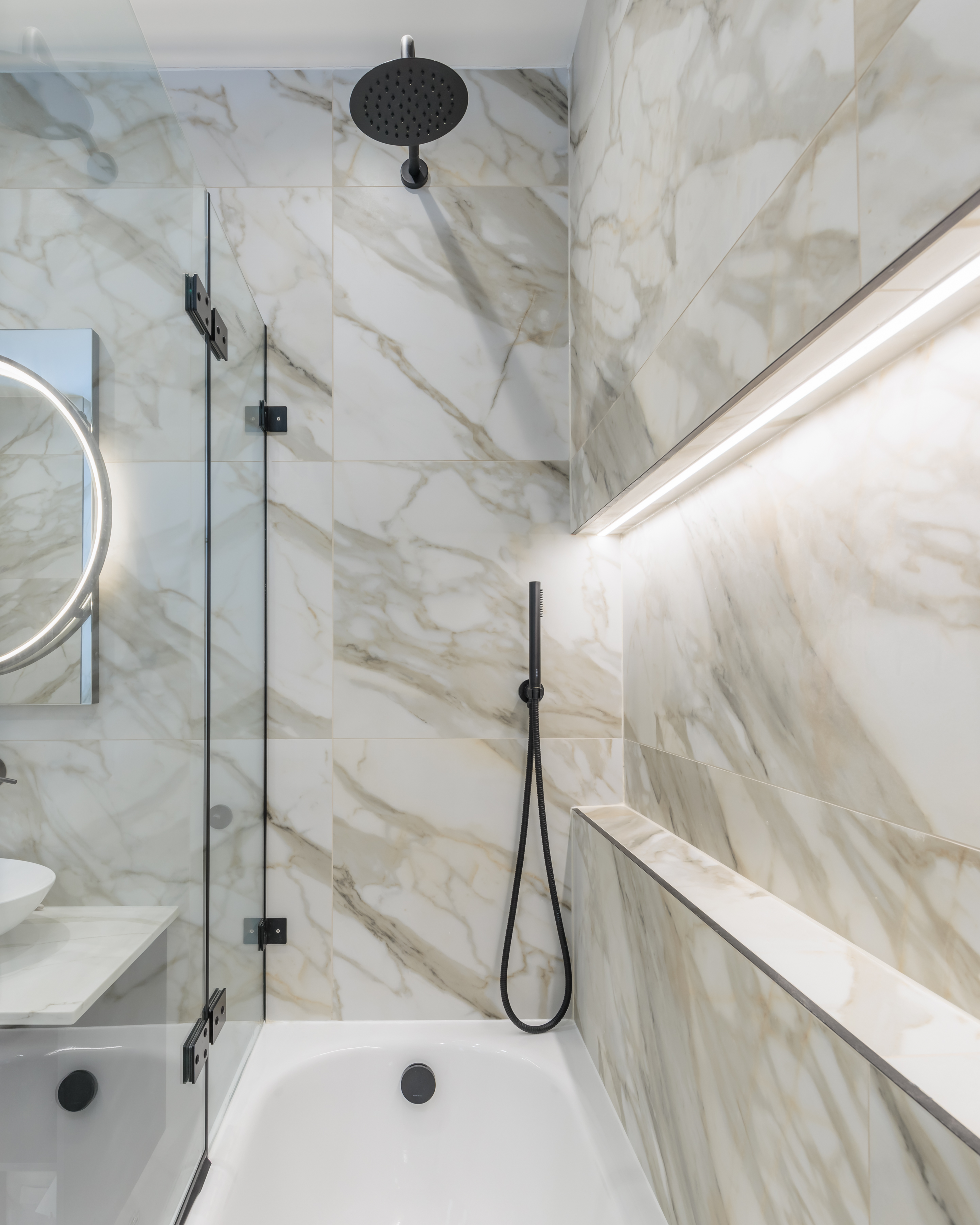 Modern bathroom featuring elegant marble wall tiles, a sleek shower with a handheld fixture, and ambient lighting. An Arch KBB team member is present, showcasing contemporary design elements.