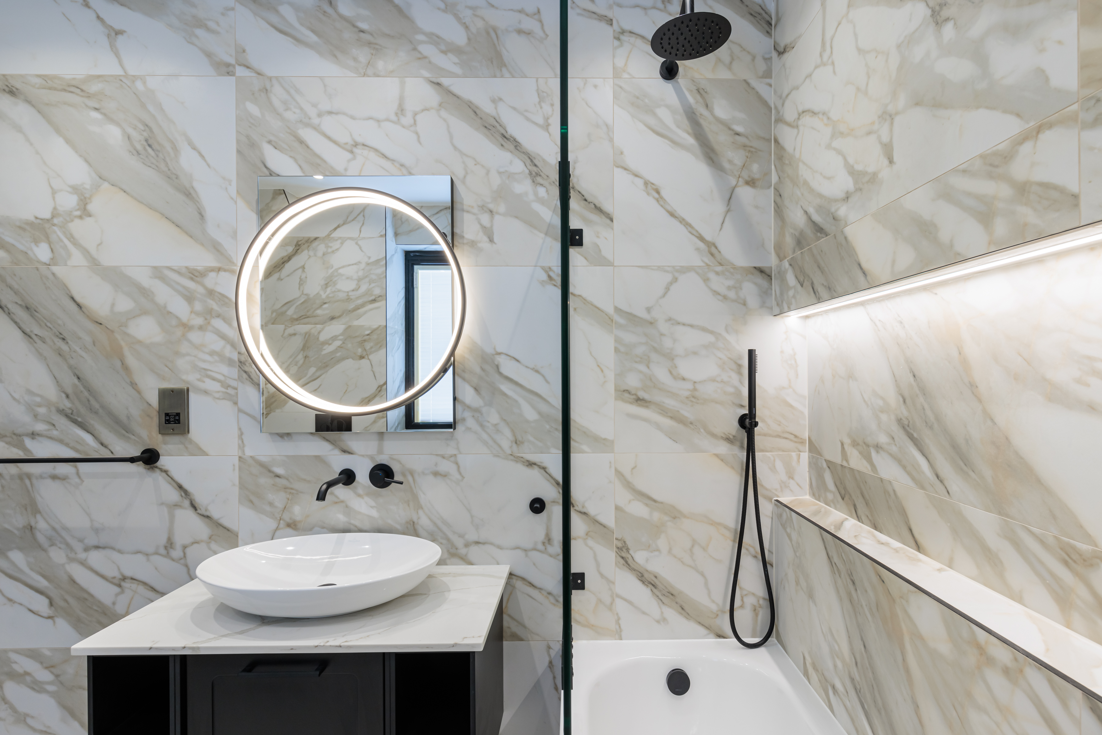 Modern bathroom design featuring marble wall tiles, a circular illuminated mirror, a stylish basin, and a sleek shower area. An Arch KBB team member is present, showcasing contemporary fixtures and elegant aesthetics.