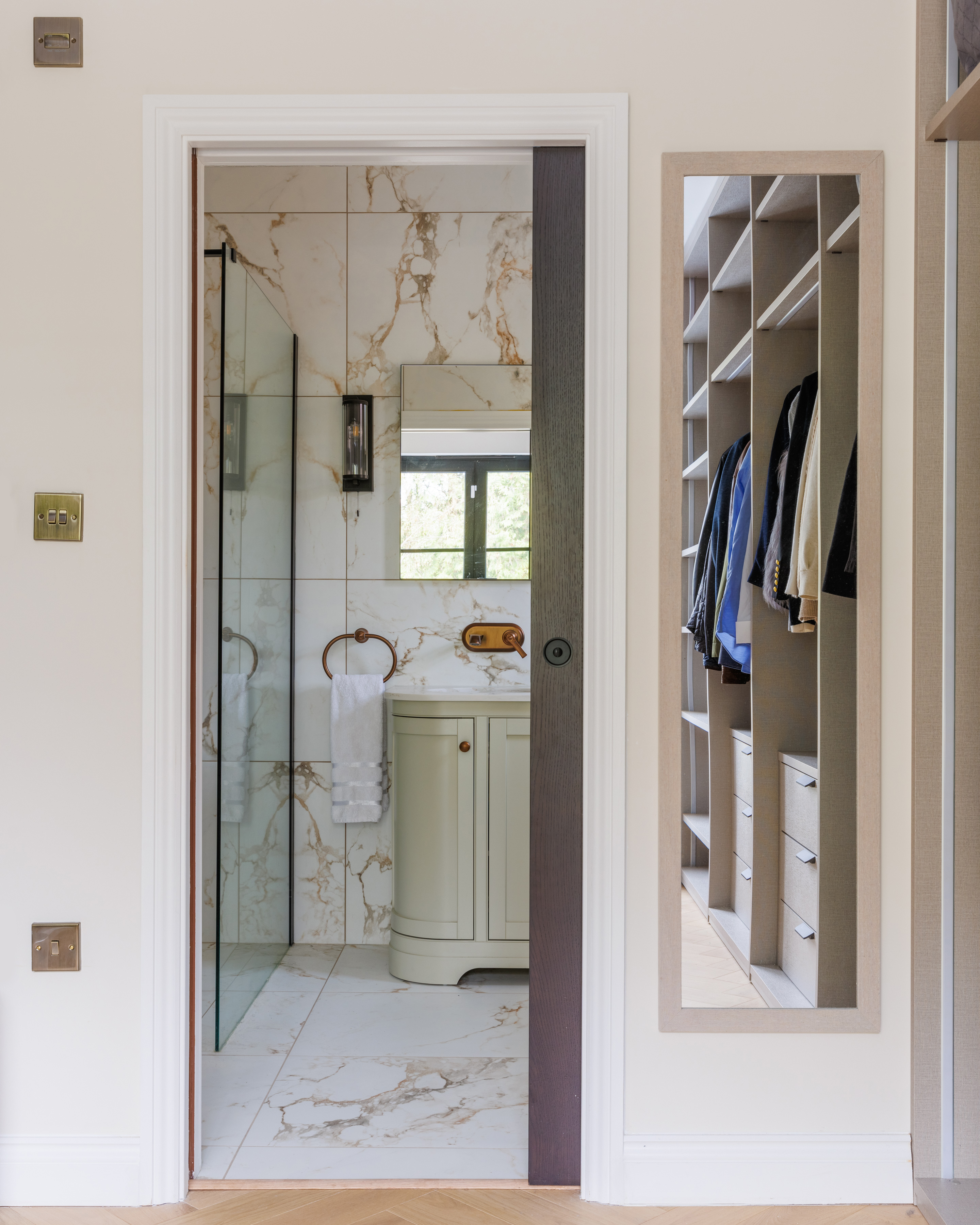 Alt text: View of a stylish bathroom featuring a glass shower enclosure, marble-patterned tiles, and a vanity unit. An Arch KBB team member is present, while a well-organised wardrobe with neatly hung clothes can be seen in the background.