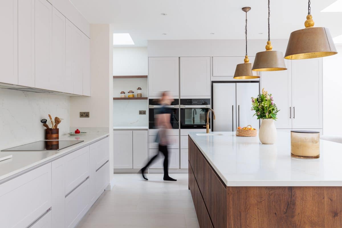 Modern kitchen featuring a large central island with a mix of white and wooden cabinetry. An Arch KBB team member is seen moving through the space, which includes stylish pendant lighting and a bouquet of flowers on the countertop. The kitchen is well-lit with natural light coming from a skylight.