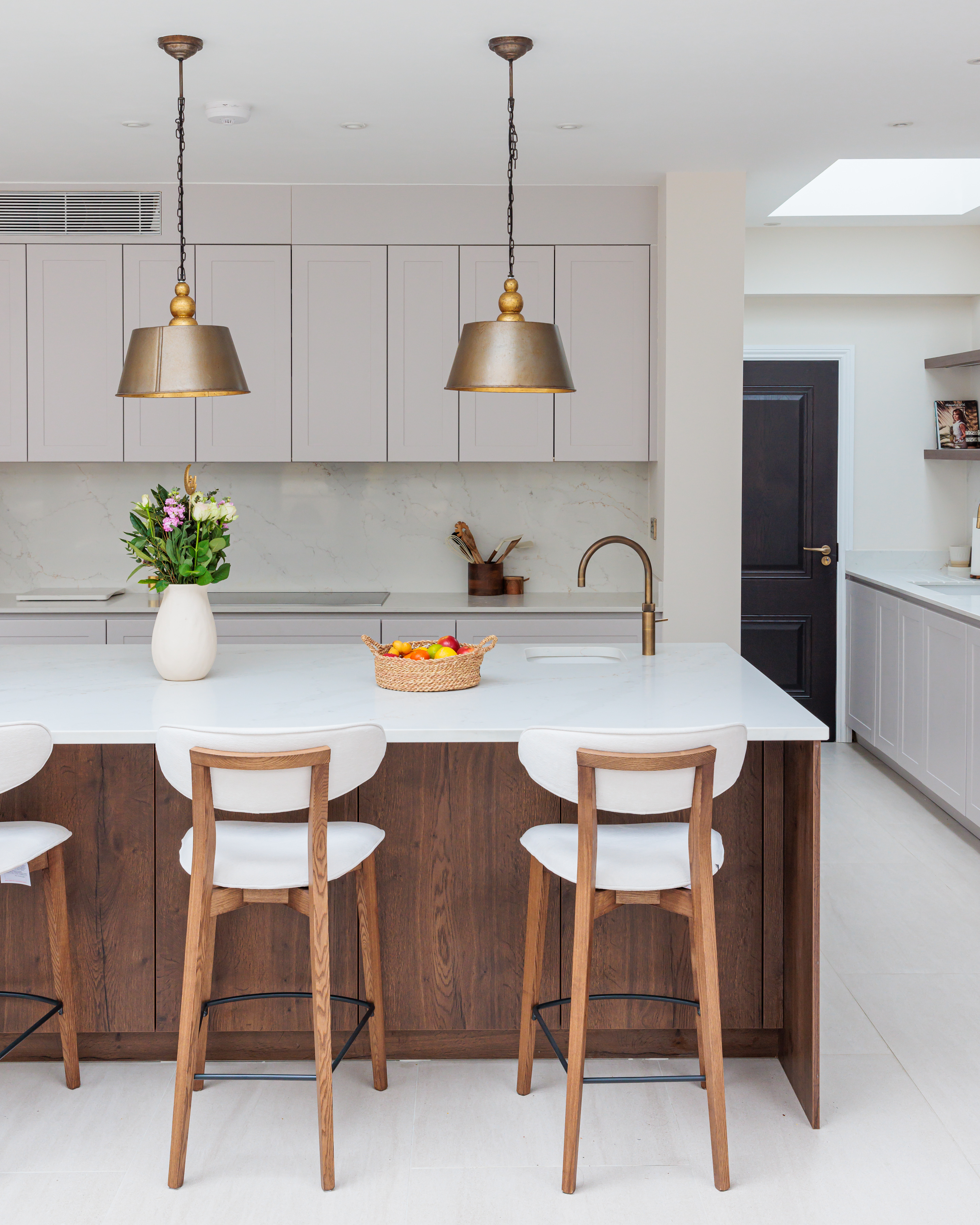 Modern kitchen interior featuring a large white island with wooden accents, three white bar stools, a vase of flowers, and a basket of fruit. Pendant lights hang above the island, illuminating the space, while sleek cabinetry and a stylish tap complete the elegant design. An Arch KBB team member is present, contributing to the inviting atmosphere.