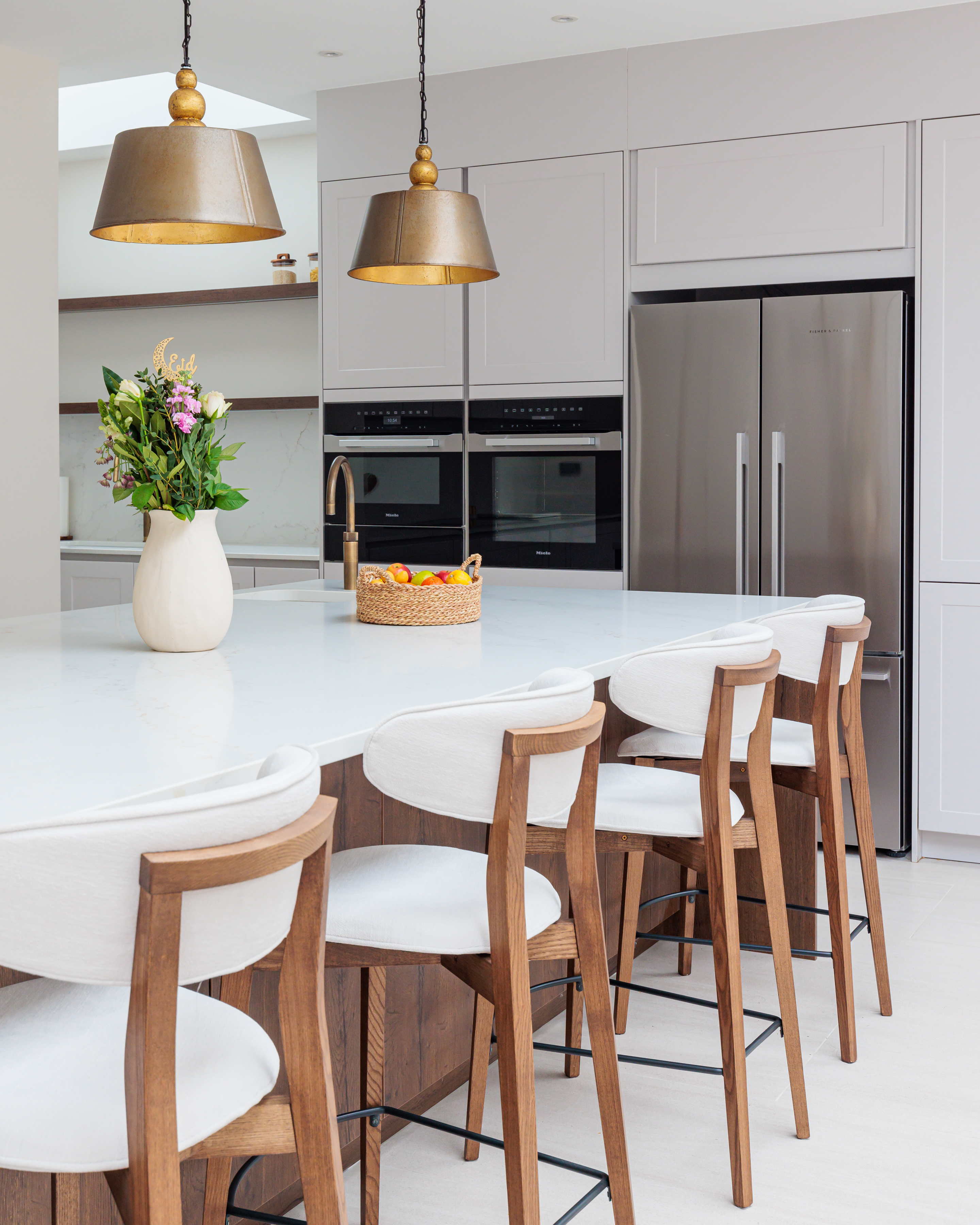 Modern kitchen featuring a spacious island with white countertop, wooden bar stools, a vase of fresh flowers, and a basket of fruit. The sleek cabinetry houses built-in ovens, and stainless steel appliances complete the contemporary design. An Arch KBB team member is present, contributing to the inviting atmosphere.