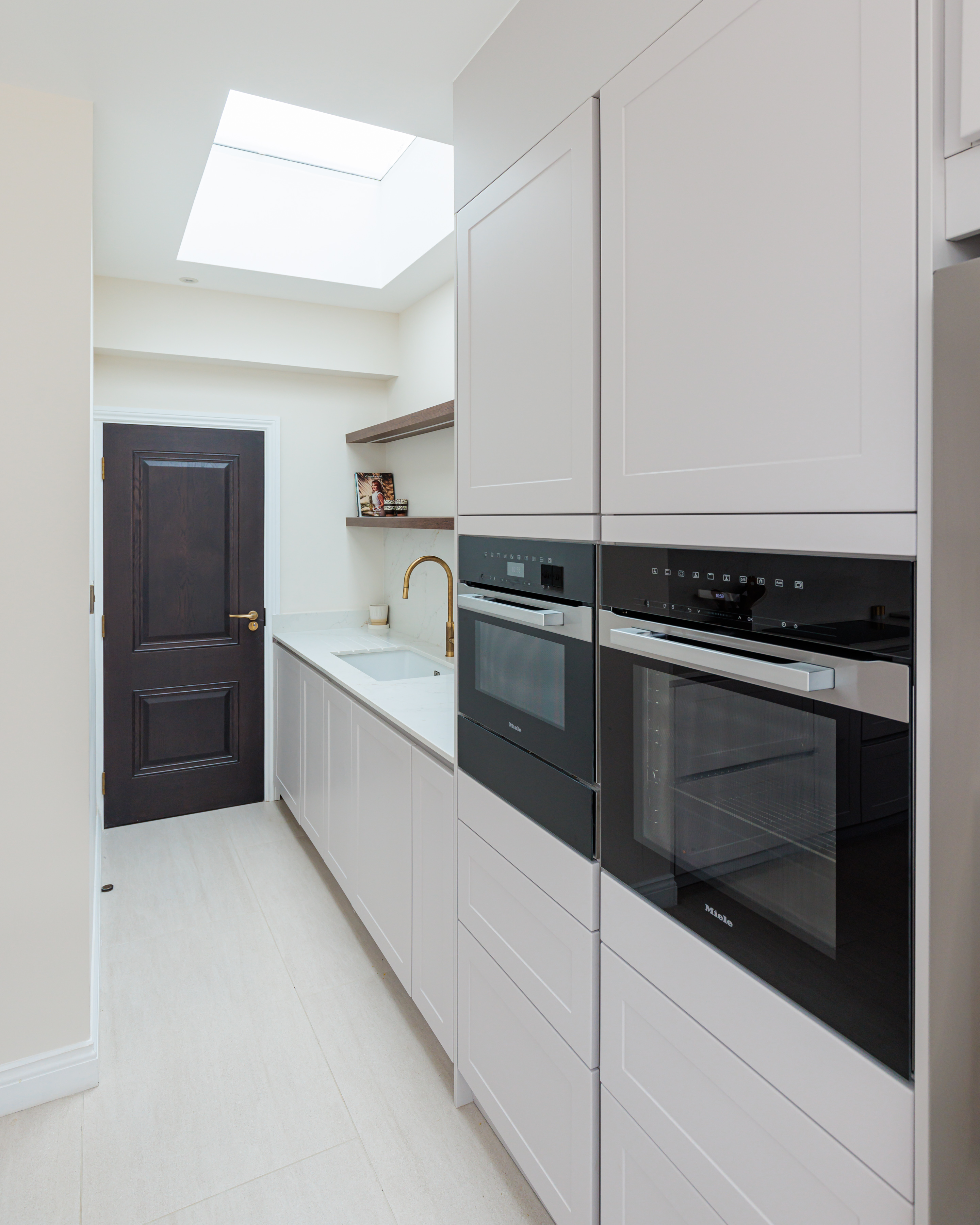 Modern kitchen interior featuring sleek cabinetry, built-in ovens, and a minimalist design. A sink and stylish tap are visible, along with a dark wooden door leading to another area. An Arch KBB team member is present, contributing to the design and functionality of the space.