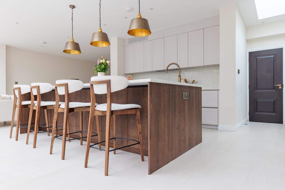 Modern kitchen interior featuring a stylish island with wooden cabinetry and seating for four. Bright pendant lights hang above the island, complementing the contemporary design. The space includes sleek cabinetry and a neutral colour palette, creating an inviting atmosphere. An Arch KBB team member is present, engaging in the kitchen environment.