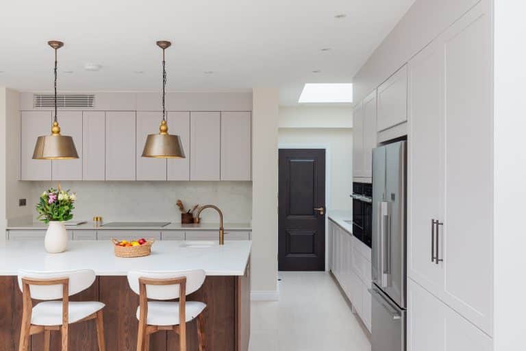 Modern kitchen interior featuring light cabinetry, a central island with seating, and elegant pendant lighting. An Arch KBB team member is present, showcasing the functional design and spacious layout. The kitchen includes stainless steel appliances and a stylish door leading to an adjoining room.