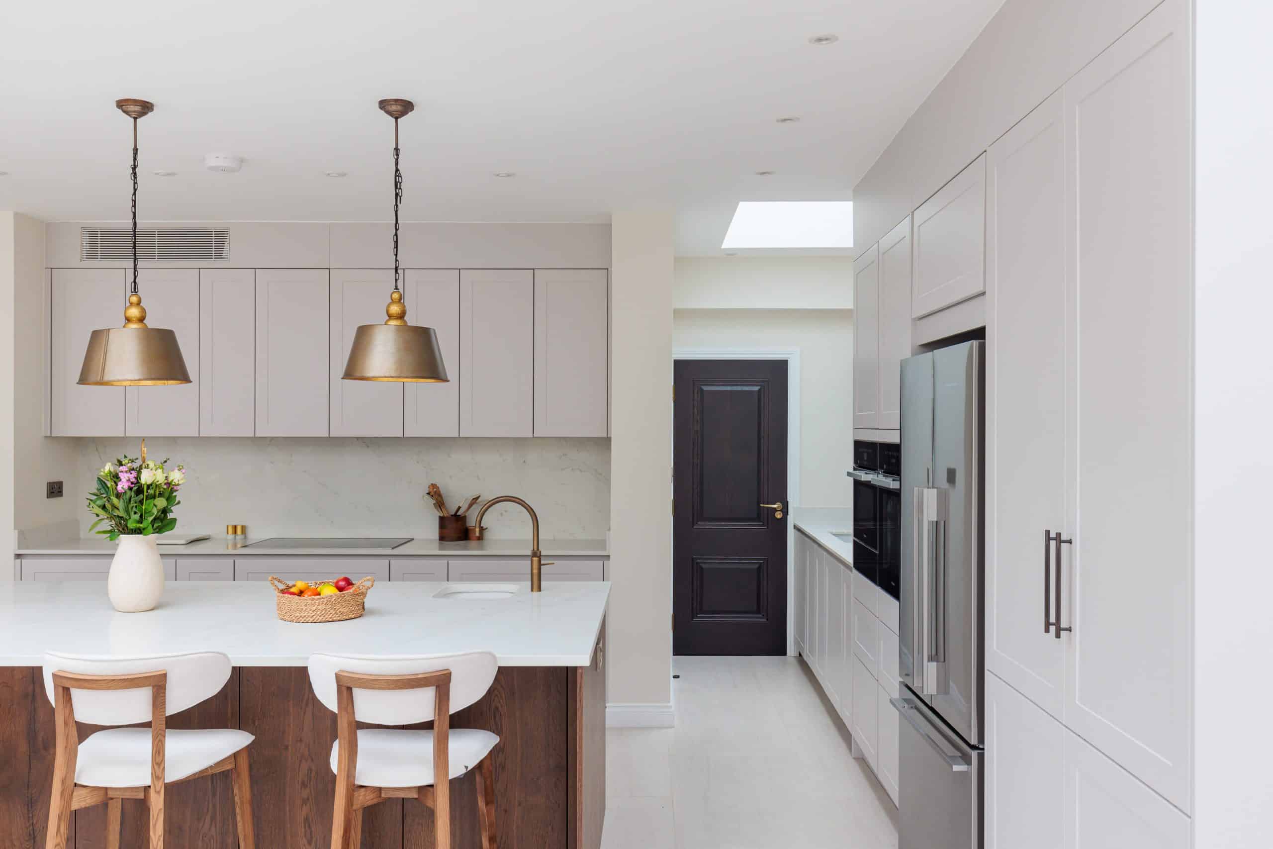 Modern kitchen interior featuring light cabinetry, a central island with seating, and elegant pendant lighting. An Arch KBB team member is present, showcasing the functional design and spacious layout. The kitchen includes stainless steel appliances and a stylish door leading to an adjoining room.