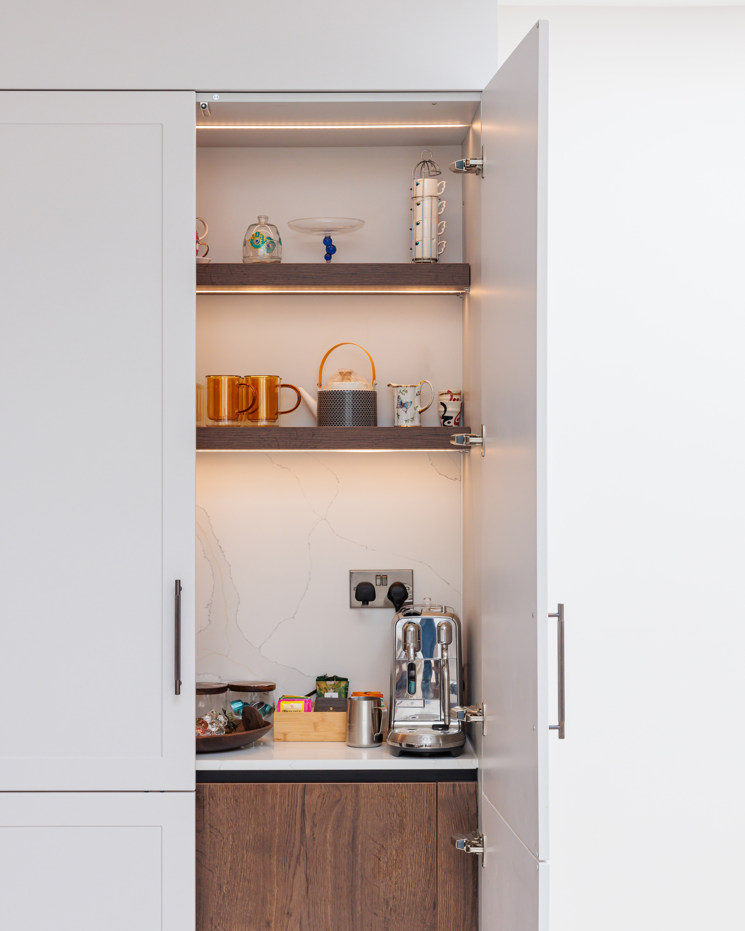 Interior view of a stylish kitchen cupboard featuring neatly arranged shelves with various mugs, decorative items, and a coffee machine, illuminated by soft lighting, showcasing modern kitchen organisation. An Arch KBB team member is present in the image.