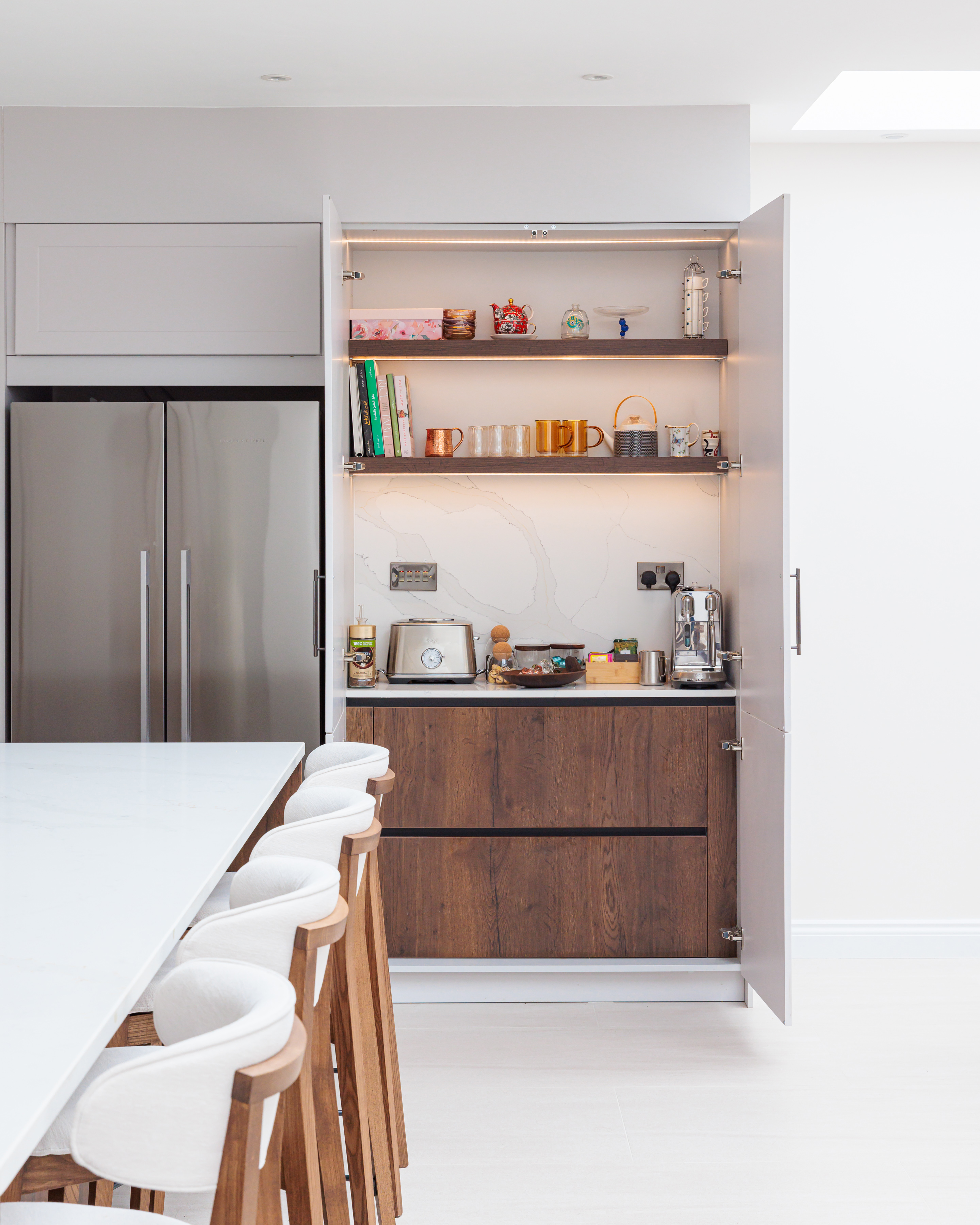 Modern kitchen interior featuring a sleek cabinetry design with open shelves displaying books, mugs, and decorative items. The kitchen includes a toaster and kettle on the countertop, complemented by a spacious island with stylish bar stools. An Arch KBB team member is present, enhancing the kitchen's inviting atmosphere.