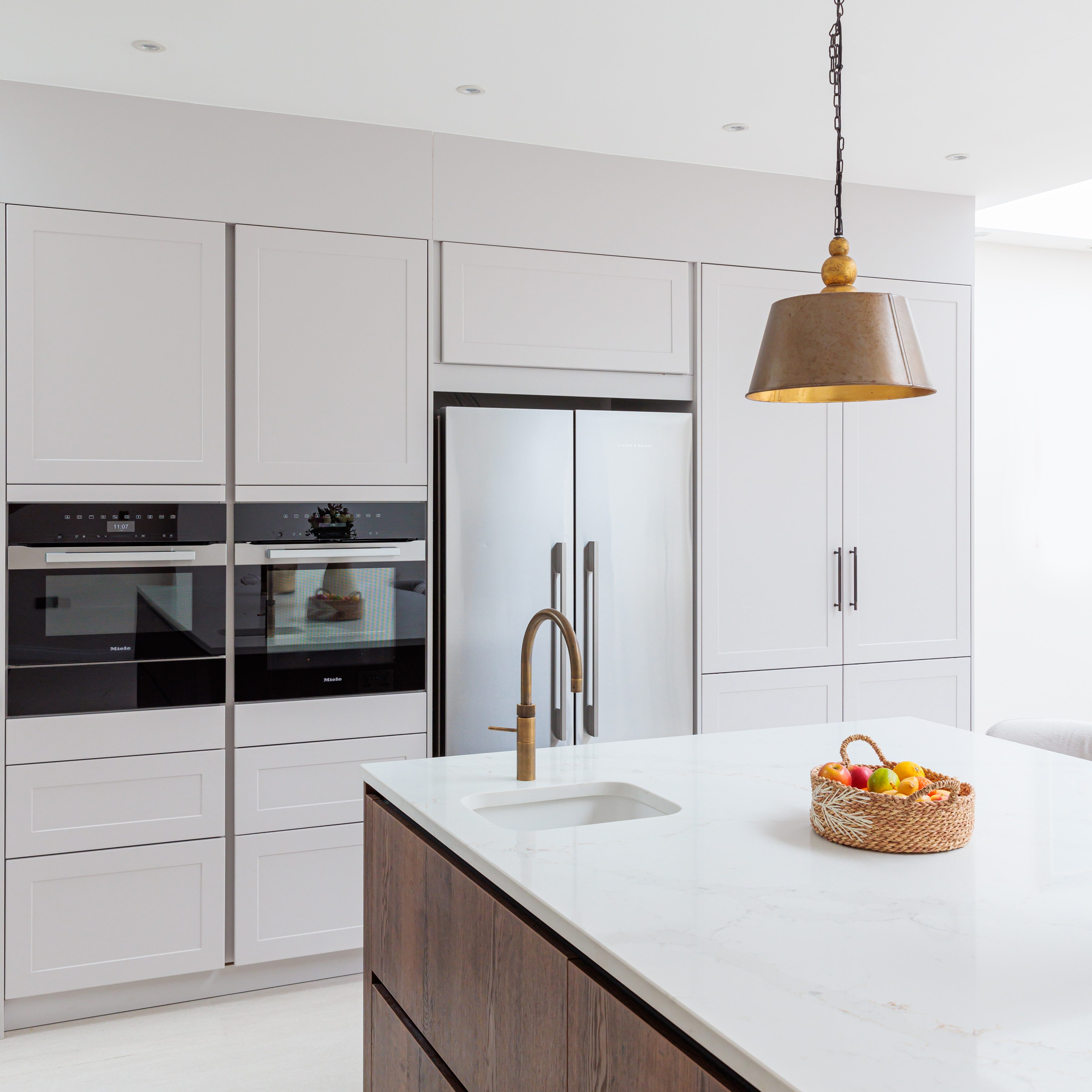 Modern kitchen featuring sleek white cabinetry, built-in appliances, and a spacious island with a sink. A stylish pendant light hangs above the island, which is adorned with a basket of fresh fruit. An Arch KBB team member is present, enhancing the kitchen's inviting atmosphere.