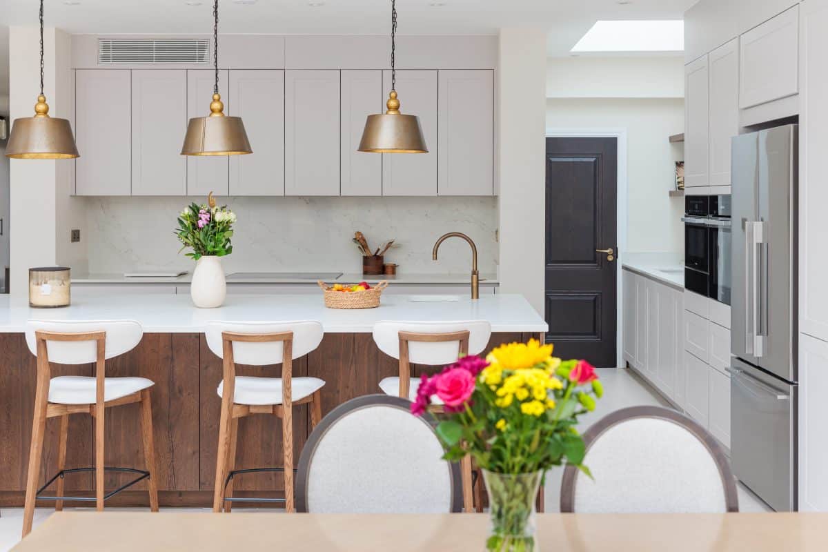 Modern kitchen interior featuring a spacious island with wooden and white cabinetry, stylish pendant lights, and a vase of fresh flowers. The design includes a dining area with a round table and additional seating at the island. An Arch KBB team member is present, contributing to the inviting atmosphere of the space.