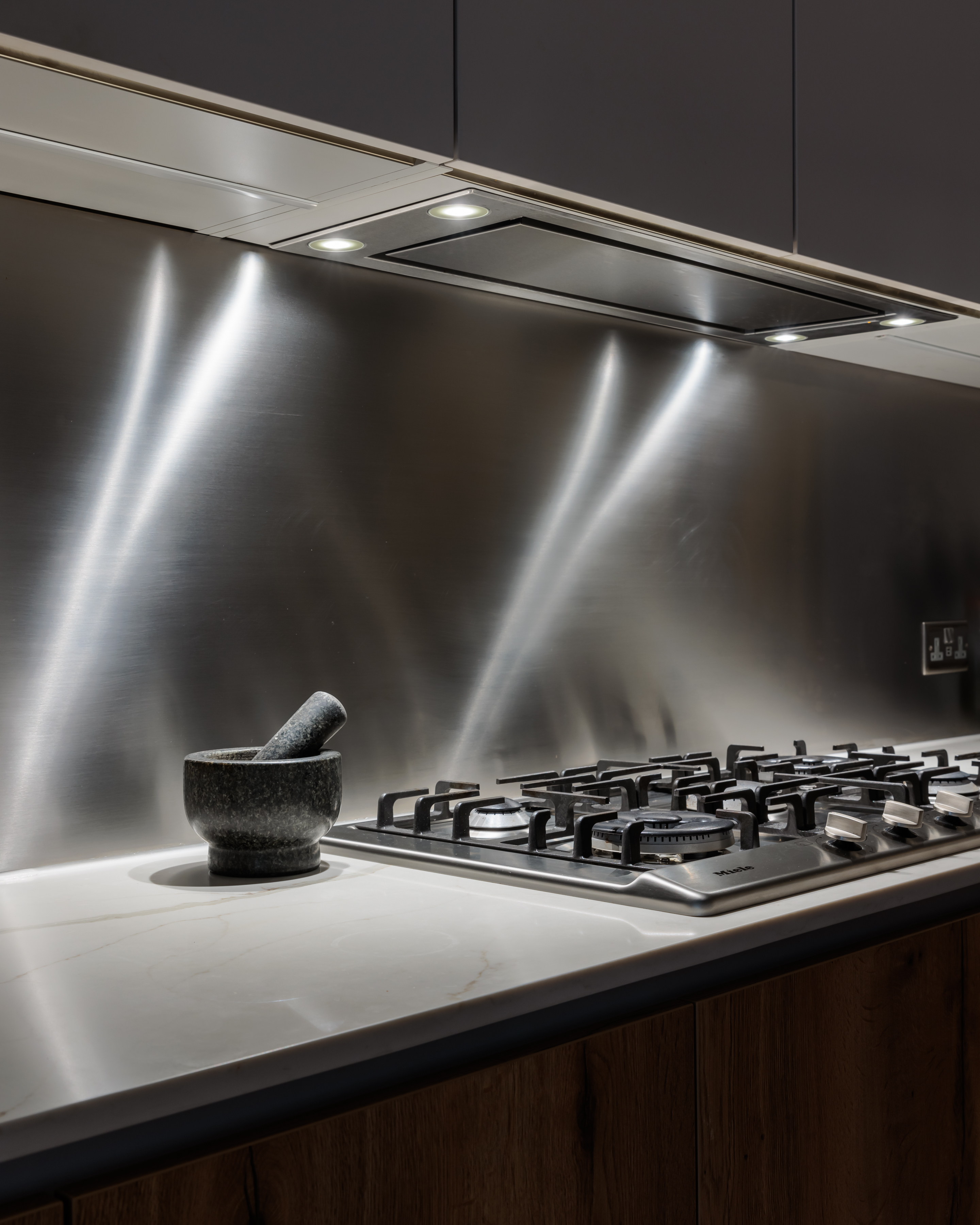 Modern kitchen featuring a sleek stainless steel backsplash with integrated lighting, a gas hob with multiple burners, and a stone mortar and pestle on the countertop, showcasing a stylish and functional cooking space designed by an Arch KBB team member.