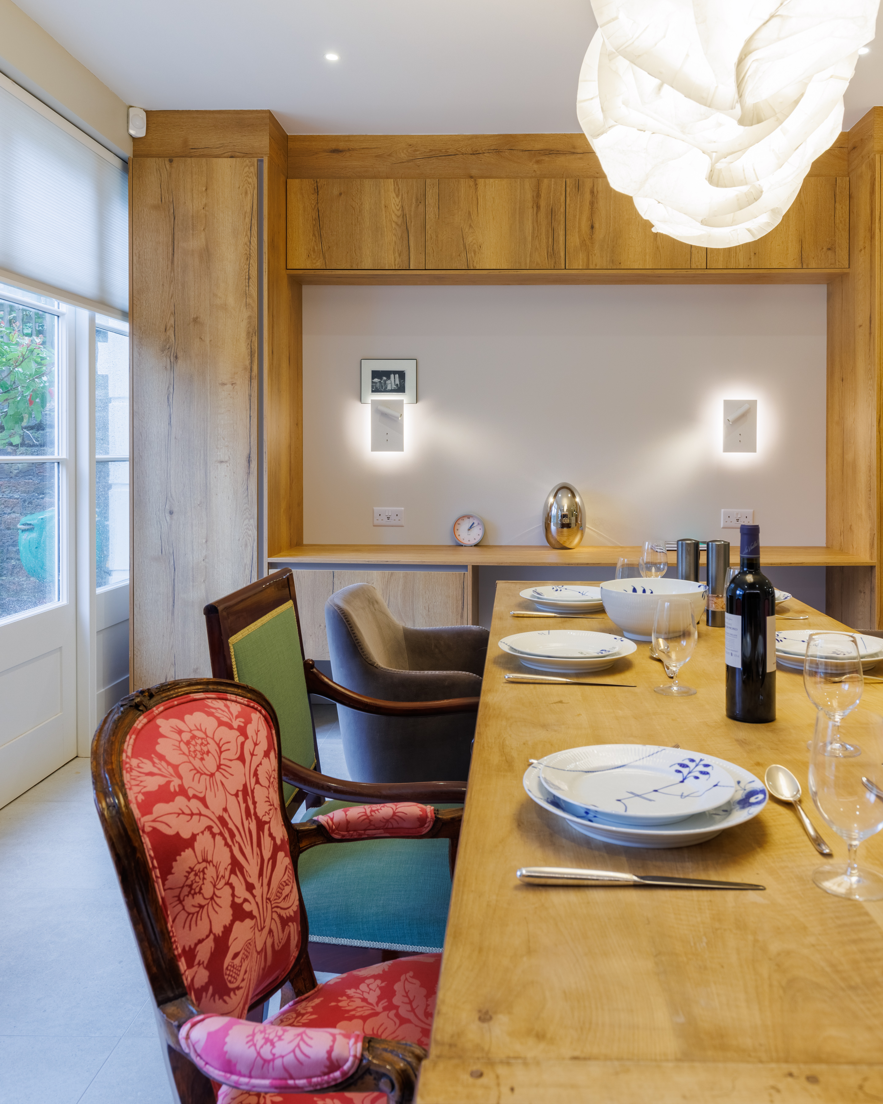 Interior view of a dining area featuring a wooden table set with plates and glasses, a bottle of wine, and a mix of modern and vintage chairs. The backdrop includes warm wooden paneling and soft lighting, creating an inviting atmosphere for dining. An Arch KBB team member is present, contributing to the design ambiance.