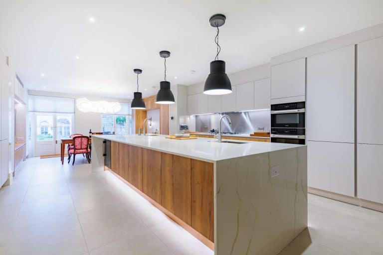 Modern kitchen featuring a spacious island with a sleek white countertop and wooden accents. The room is illuminated by stylish pendant lights and includes integrated appliances. An Arch KBB team member is present, adding to the design-focused atmosphere. The overall aesthetic is contemporary and inviting, perfect for both cooking and entertaining.