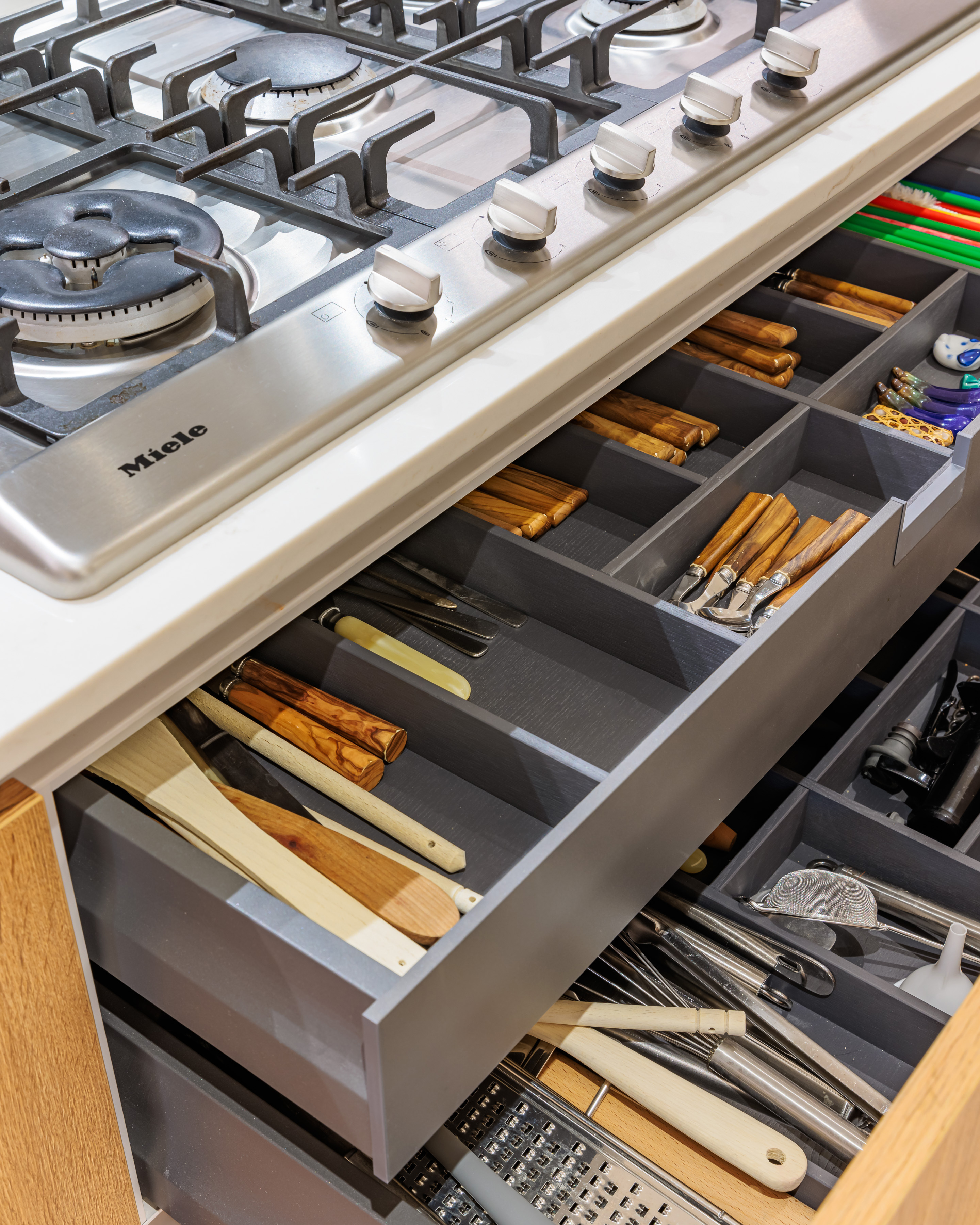 Image of a modern kitchen featuring a Miele gas hob and an organised drawer filled with various kitchen utensils and tools. The drawer showcases wooden-handled utensils, knives, and other cooking implements, highlighting efficient kitchen storage solutions. An Arch KBB team member is present, demonstrating the kitchen's functionality.