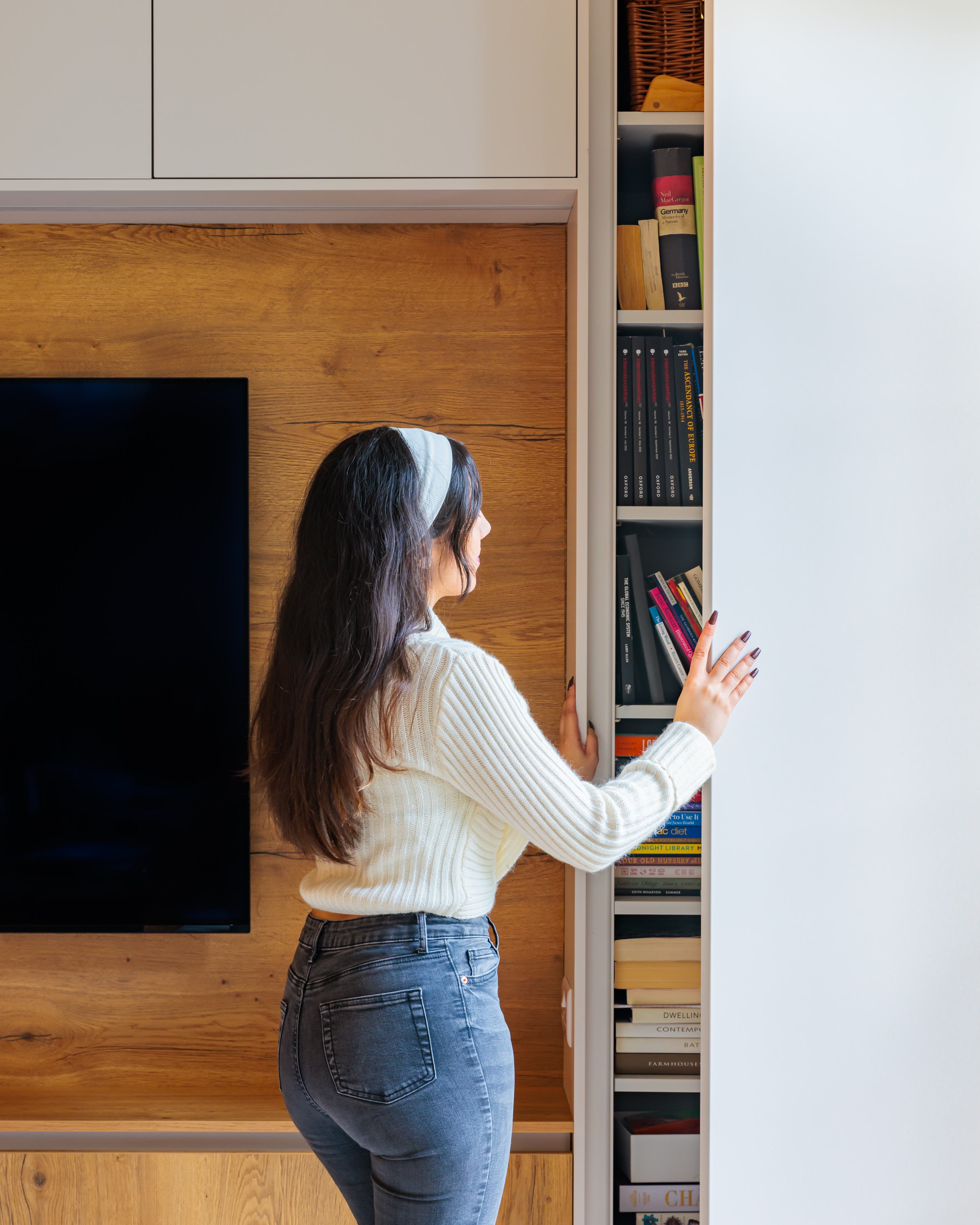 Arch KBB team member interacting with a hidden bookshelf in a modern living space, featuring a large television on a wooden wall. The bookshelf contains various books, showcasing a blend of style and functionality in interior design.