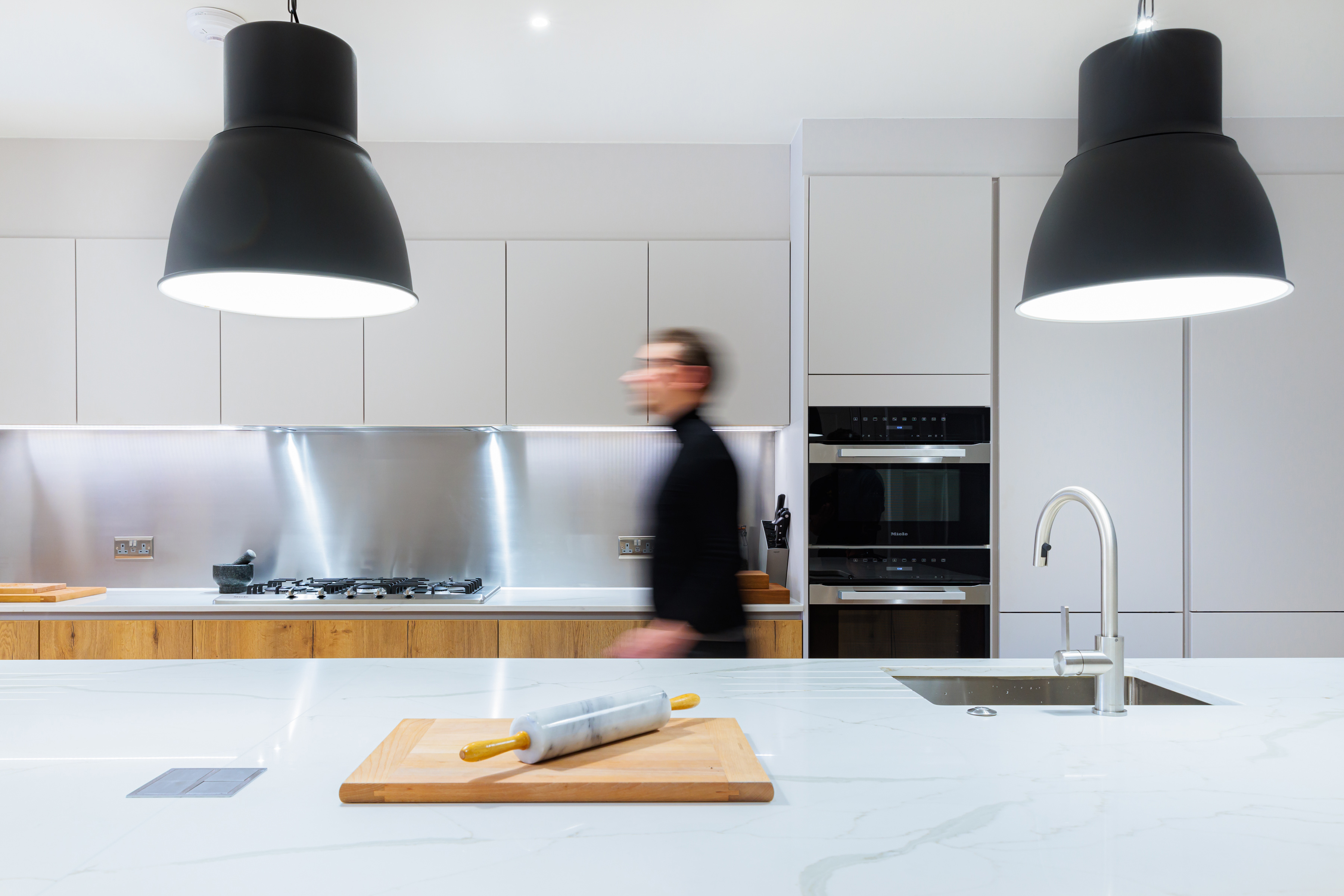 Arch KBB team member in a modern kitchen with a marble countertop and wooden cabinetry, featuring a rolling pin on a chopping board, and stylish pendant lighting above. The background includes a stainless steel backsplash and kitchen appliances.