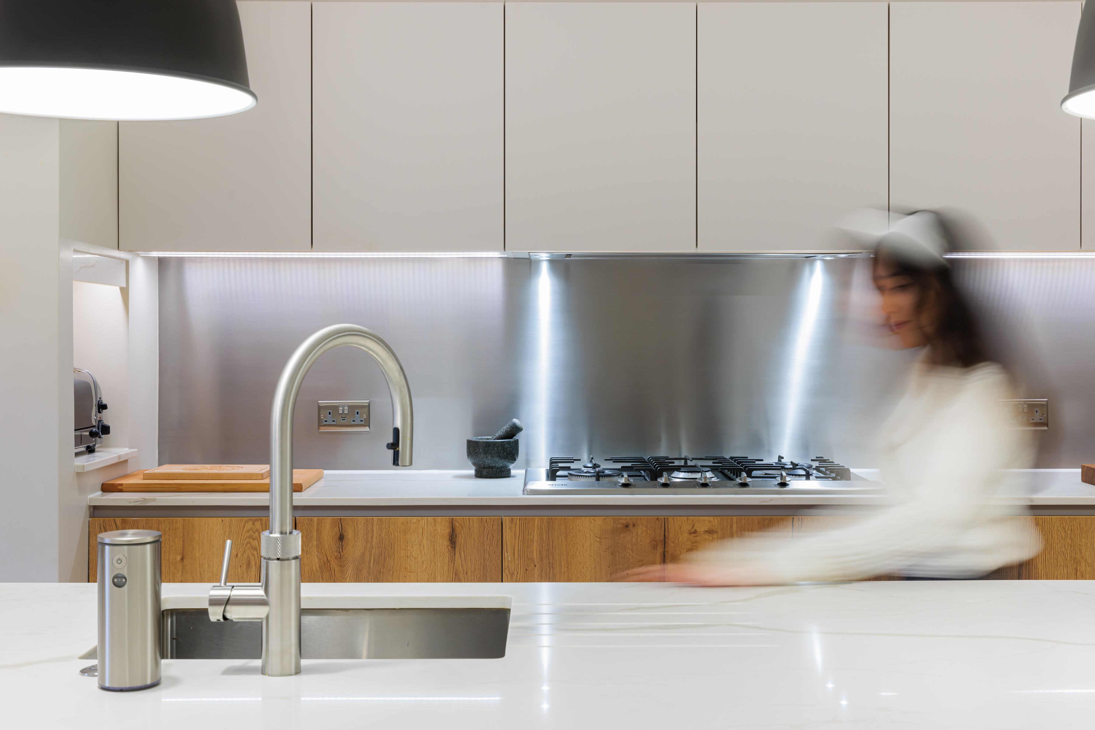A modern kitchen featuring a sleek stainless steel backsplash, contemporary cabinetry, and a stylish sink. An Arch KBB team member is engaged in kitchen activities, enhancing the space's functionality and design.