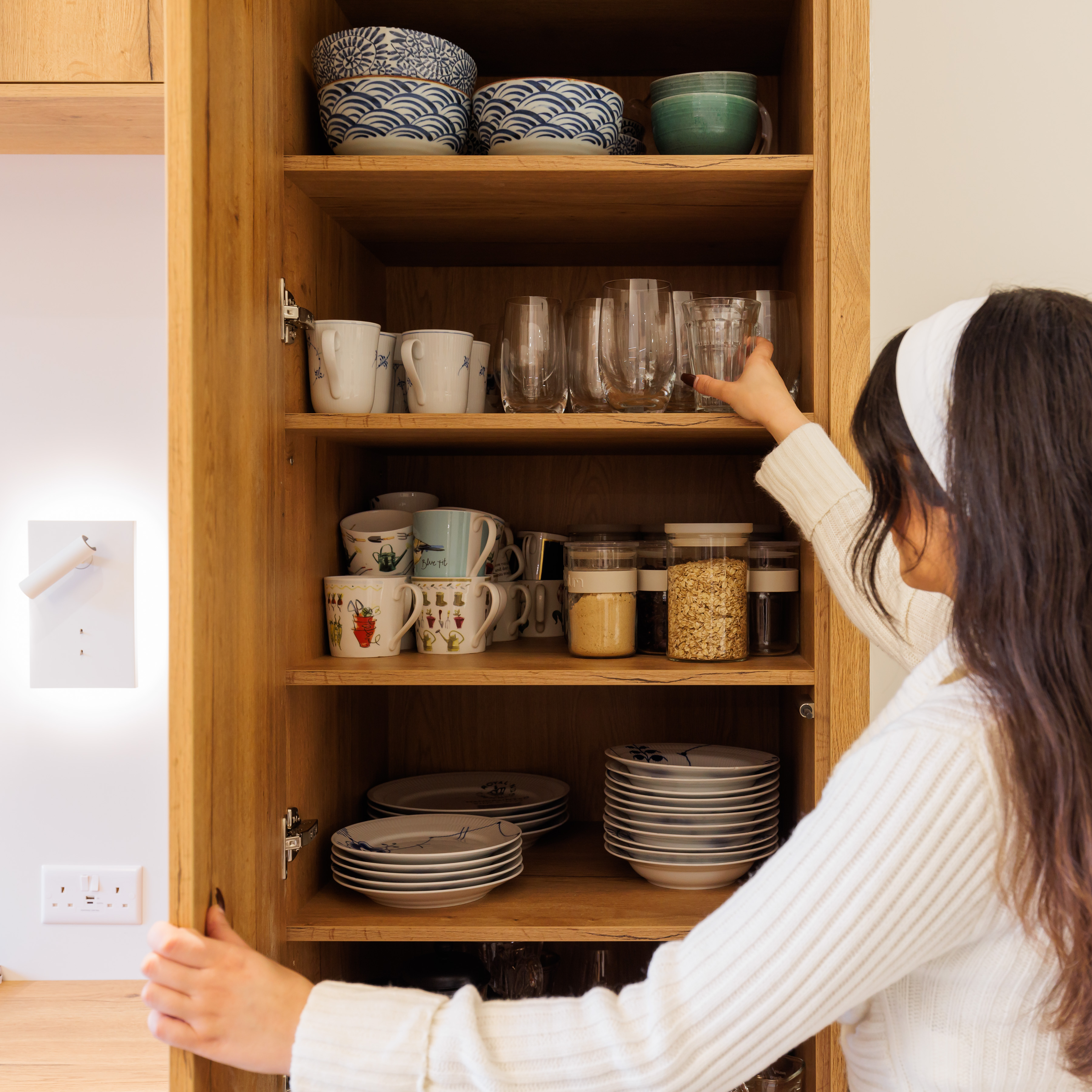 Arch KBB team member organising kitchen cupboard with various glassware, mugs, and storage jars, showcasing a neat arrangement of dinnerware and utensils.