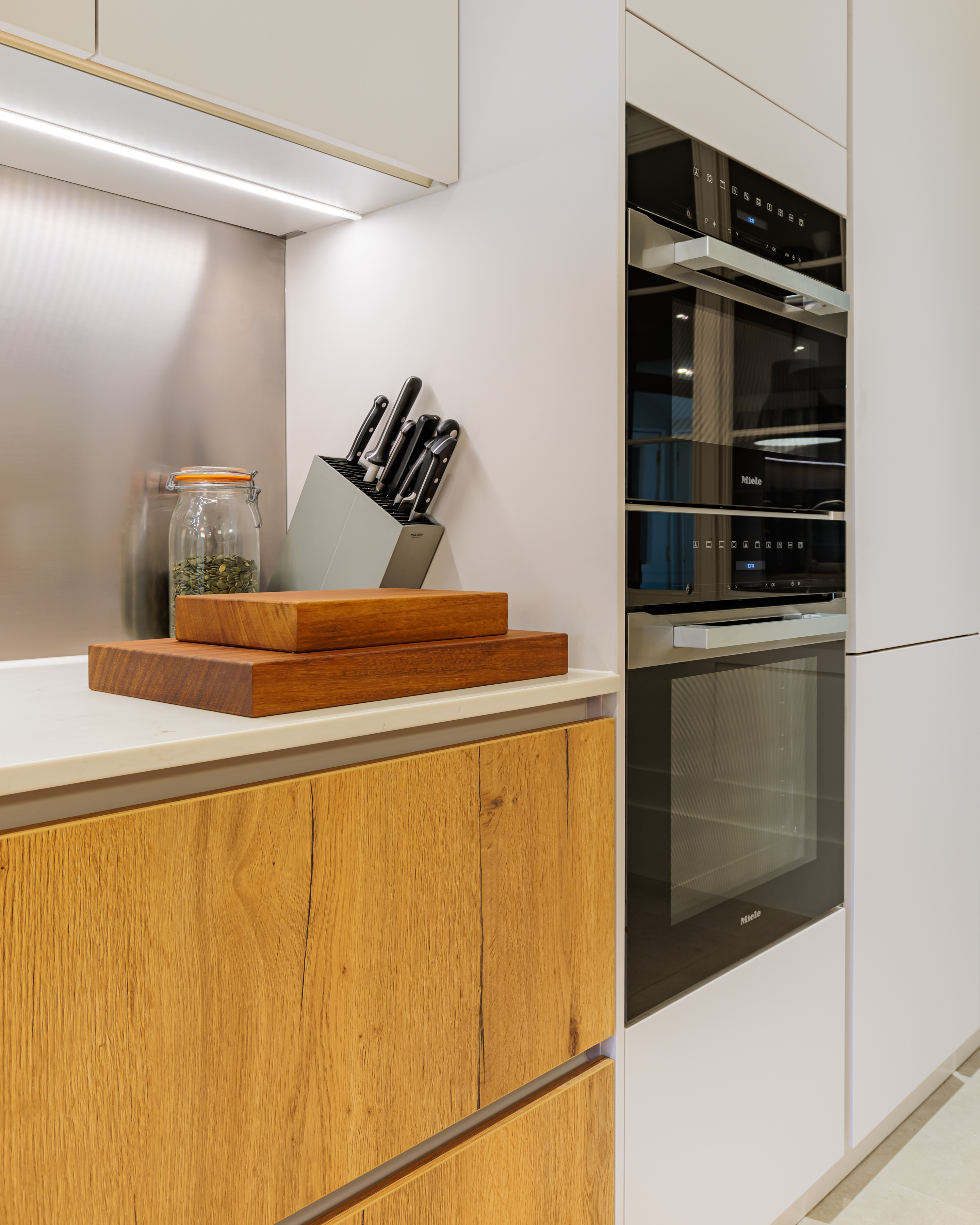 Modern kitchen interior featuring a sleek countertop with wooden chopping boards, a knife block, and a glass jar of herbs. The cabinetry combines a light finish with natural wood grain, complemented by built-in appliances for a contemporary look. An Arch KBB team member is present, enhancing the design atmosphere.