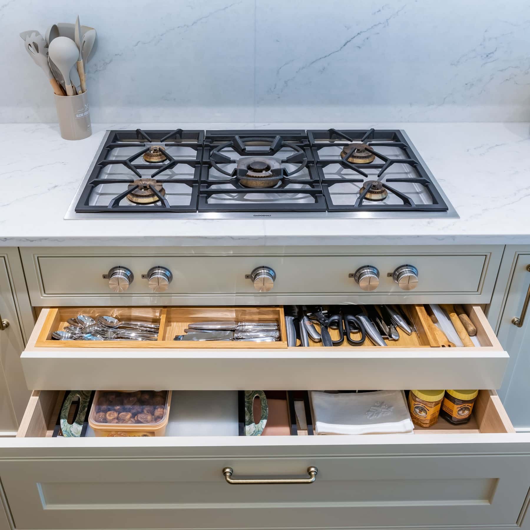 A well-organised kitchen featuring a gas hob on a marble countertop, with a drawer open to reveal neatly arranged cutlery and kitchen utensils. An Arch KBB team member is present, showcasing the functional design and storage solutions in modern kitchen spaces.