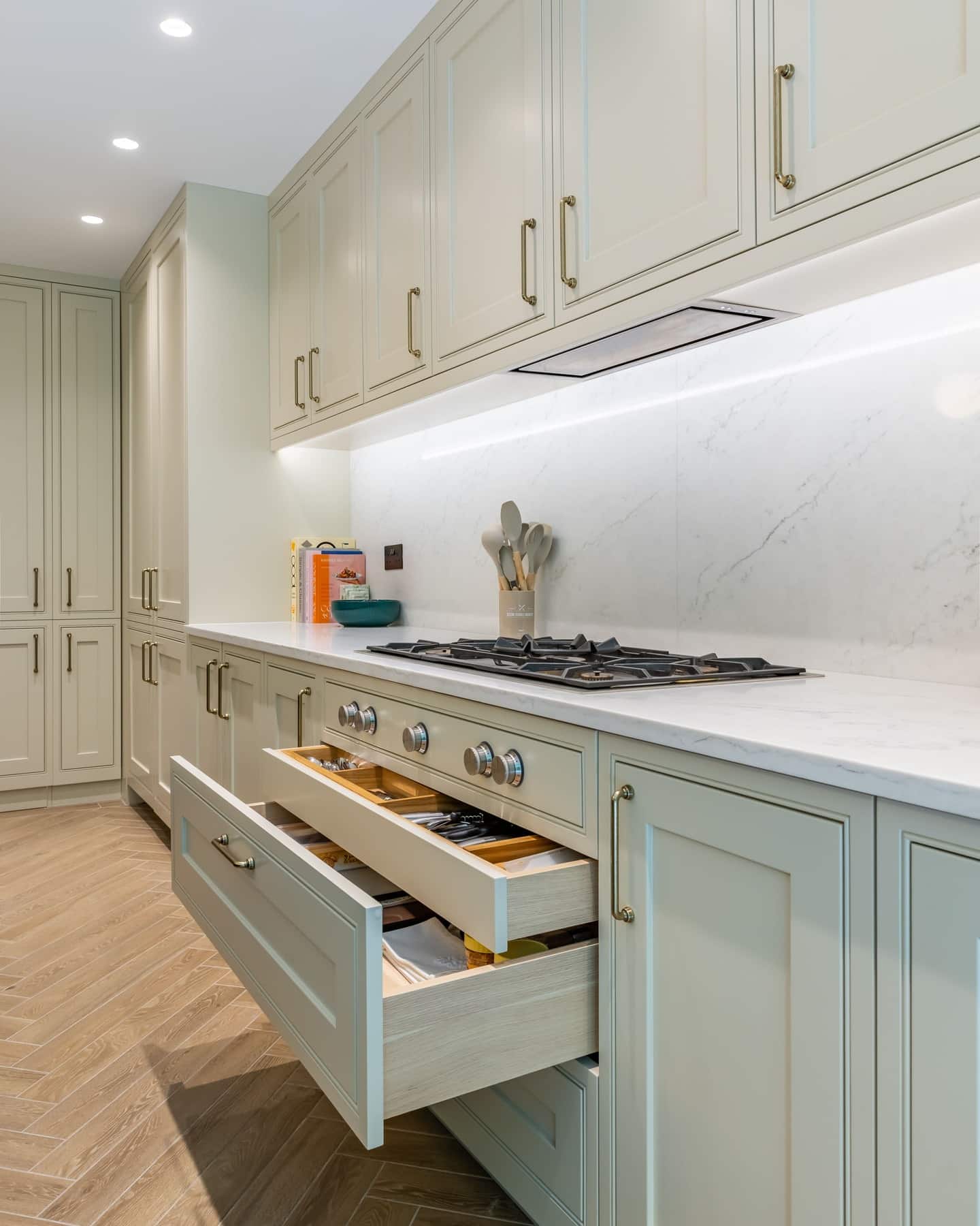 Modern kitchen featuring a sleek design with light green cabinetry, a marble countertop, and a gas hob. Open drawer displaying utensils, complemented by a minimalist backsplash and integrated lighting. An Arch KBB team member is present, enhancing the atmosphere of the stylish kitchen space.