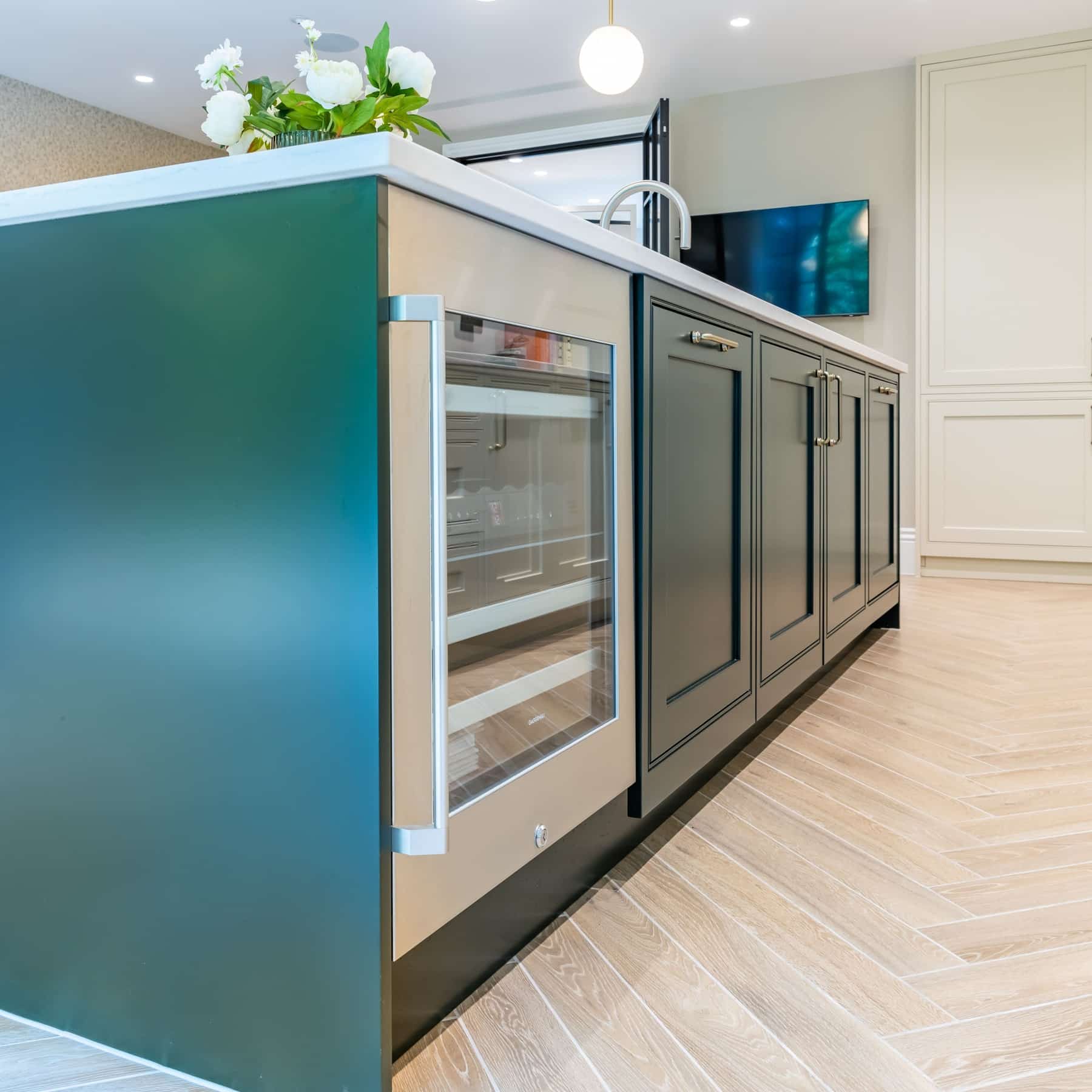 Modern kitchen featuring a stylish island with dark cabinetry and a stainless steel wine fridge. An Arch KBB team member is visible in the background, contributing to the design. The flooring is elegantly laid in a herringbone pattern, enhancing the contemporary aesthetic.