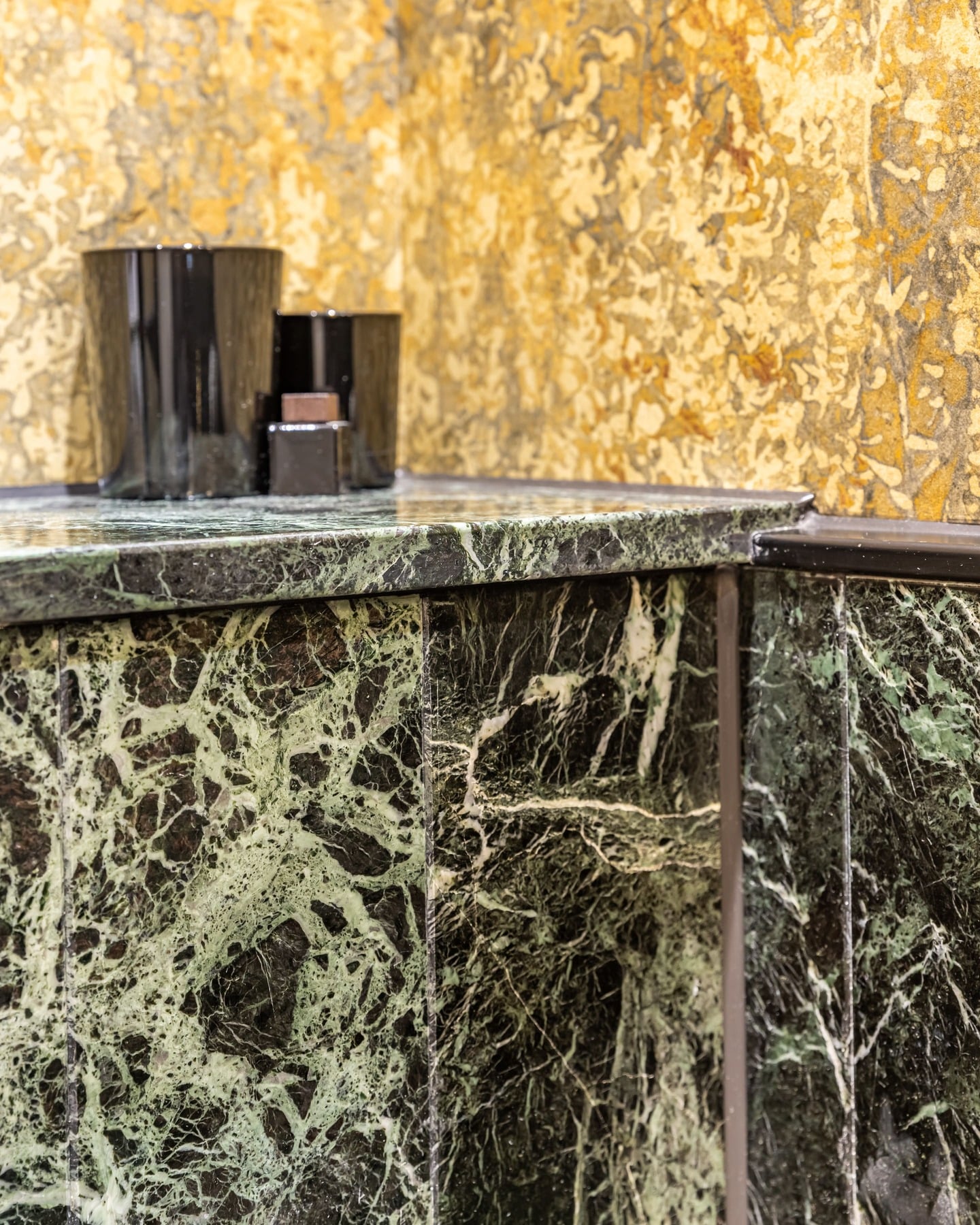 Close-up of a luxurious green marble countertop featuring intricate veining, complemented by a textured wall and stylish black accessories. An Arch KBB team member is present, showcasing modern design elements.
