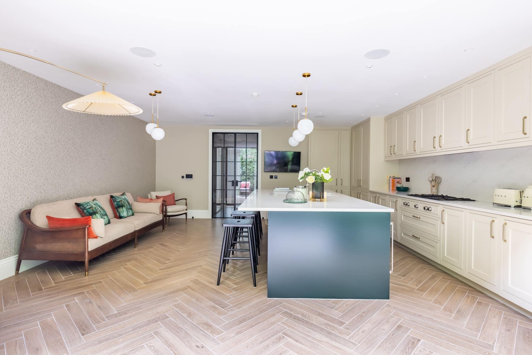 Modern kitchen and living area featuring a stylish open-plan design. The space includes a large kitchen island with seating, elegant cabinetry, and a comfortable sofa with decorative cushions. Pendant lighting adds a warm touch, while the herringbone floor pattern enhances the contemporary aesthetic. An Arch KBB team member is present, showcasing the inviting atmosphere of the room.