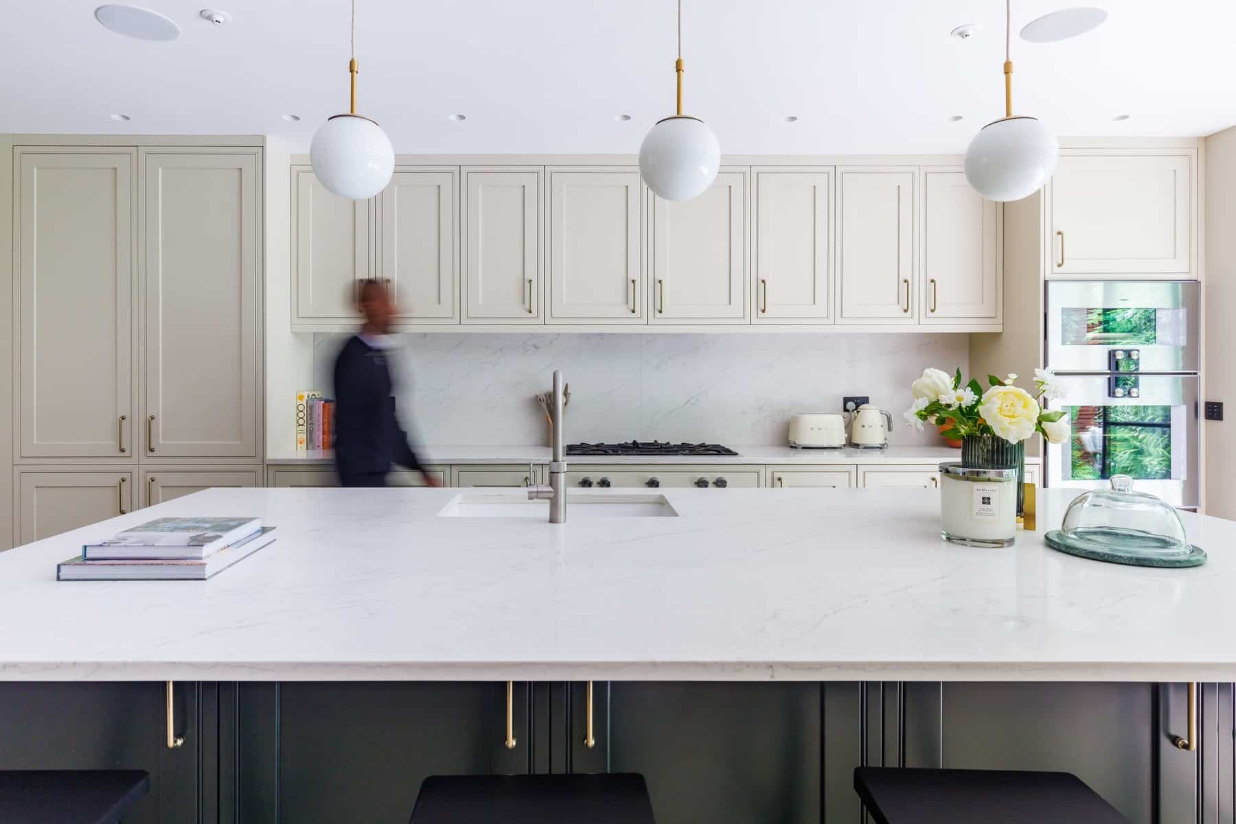 Modern kitchen featuring a spacious marble island, sleek cabinetry, and pendant lighting. An Arch KBB team member is seen in motion, adding a dynamic element to the contemporary design. Fresh flowers and kitchen appliances complete the stylish interior.