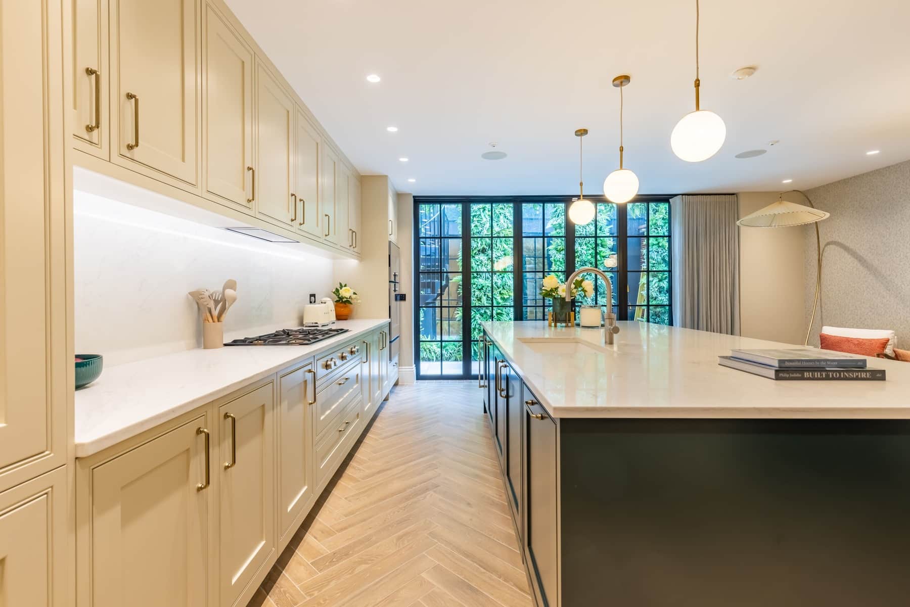 Modern kitchen interior featuring a spacious layout with light-coloured cabinetry, a large central island, and stylish pendant lighting. The space includes a gas stove and a view of greenery through large glass doors, enhancing natural light. An Arch KBB team member is present, showcasing the design in a contemporary home setting.