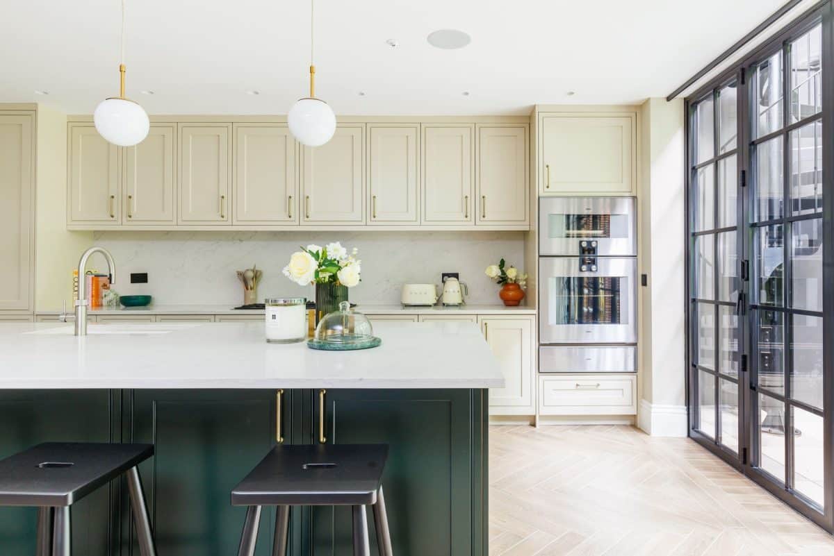 Modern kitchen featuring a stylish island with black stools, cream cabinetry, and a sleek stainless steel refrigerator. A vase of white roses adds a touch of elegance, while large windows allow natural light to illuminate the space. An Arch KBB team member is present, contributing to the design ambiance.