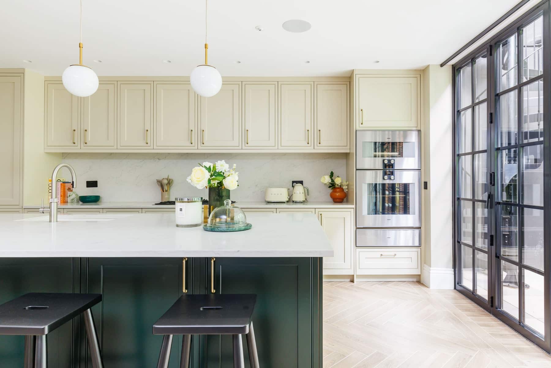 Modern kitchen featuring a stylish island with black stools, cream cabinetry, and a sleek stainless steel refrigerator. A vase of white roses adds a touch of elegance, while large windows allow natural light to illuminate the space. An Arch KBB team member is present, contributing to the design ambiance.