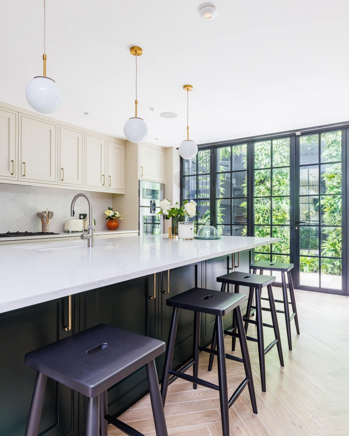 Modern kitchen design featuring a spacious marble island with black stools, stylish cabinetry, and large windows that overlook a lush garden. An Arch KBB team member is present, enhancing the inviting atmosphere.