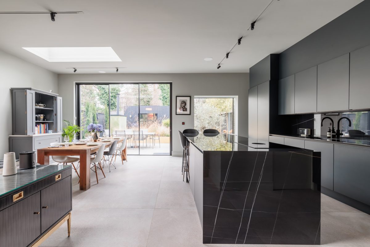 Modern kitchen and dining area featuring sleek black cabinetry, a large island with a marbled countertop, and a spacious dining table. Natural light floods the space through large glass doors leading to an outdoor area, complemented by minimalist decor and stylish furnishings.